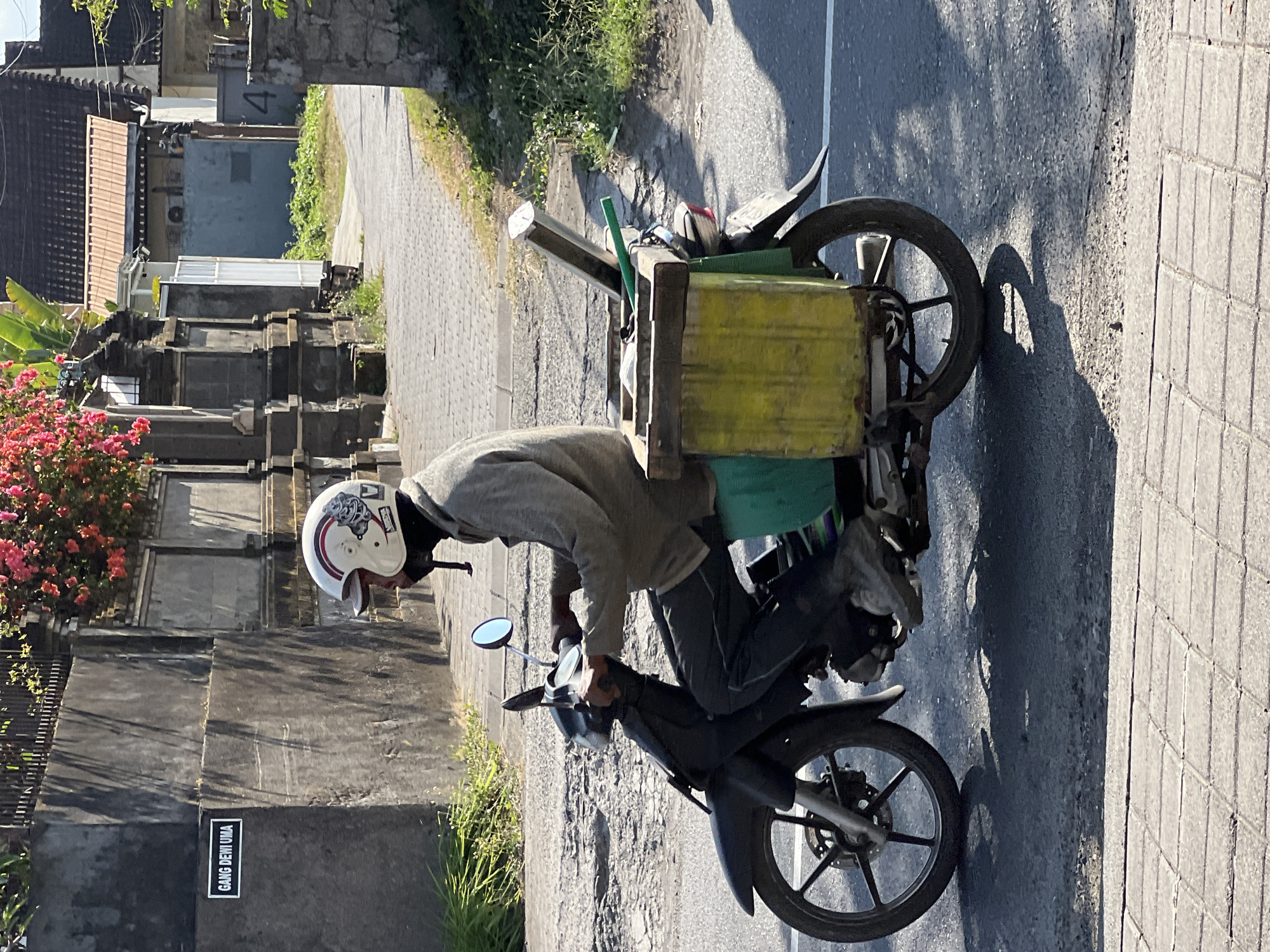 A person wearing a white and red helmet rides a black motorcycle with a yellow and green cargo box attached to the rear. The motorcycle is in motion on a paved road, casting a shadow. The person is wearing a light-colored long-sleeved shirt and dark pants. The cargo box appears to hold some items, including what looks like a green pole. In the background, a gray wall with a sign that reads "GANG DEWI UMA" is visible, along with stone pillars and lush green vegetation. Buildings with tiled roofs are also present. The scene is set in Denpasar, Indonesia, indicated by the street sign.
