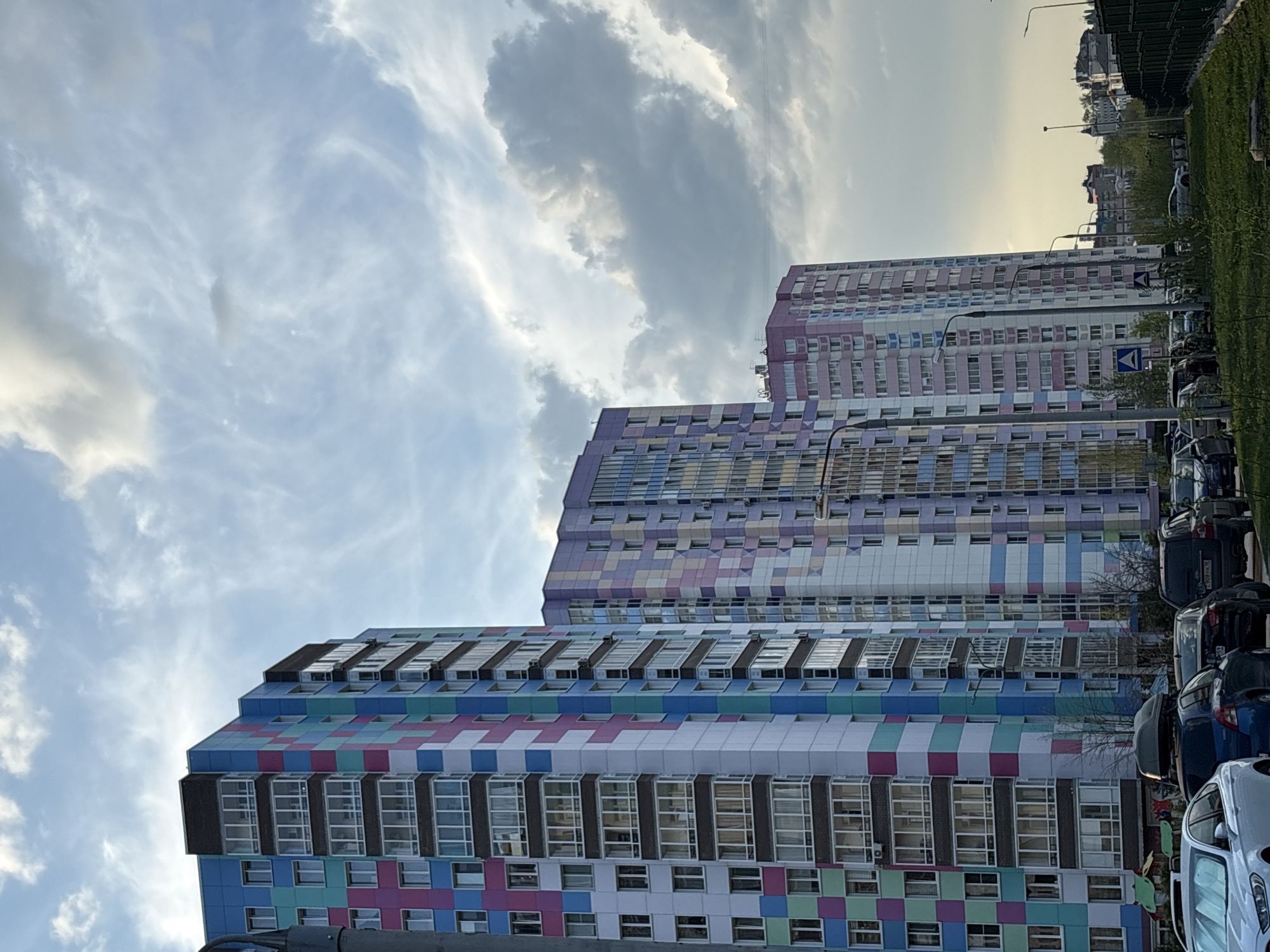The image shows three tall, colorful apartment buildings in Perm, Russia. The buildings are decorated with patterns of squares in blue, pink, green, white, and purple. Windows are visible on each building level. The sky overhead is partly cloudy with a mix of white and gray clouds. Cars are parked along the side of the road near the buildings. Green grass and sparse trees are seen near the top-right edge of the image, with other buildings visible in the distance. The photograph is taken from a low angle, emphasizing the height of the apartment buildings.