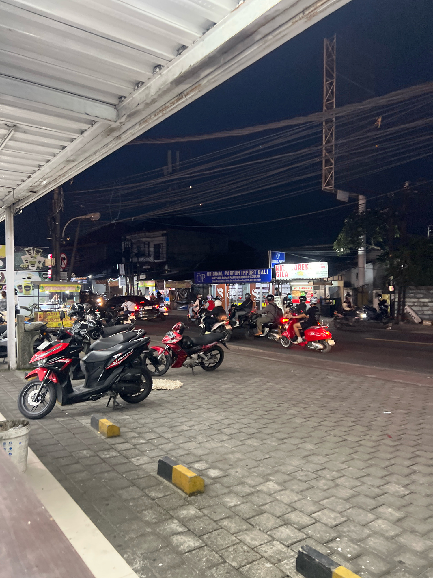 This is a nighttime street scene in Denpasar, Indonesia, likely an urban commercial area. A covered walkway with a corrugated metal roof runs along the left side of the image, providing shelter over a paved sidewalk. On the sidewalk, several motorcycles are parked, with two prominent scooters in the foreground: one red and black, and another red.
The street itself is paved and busy with traffic. Multiple motorcycles, many with riders wearing helmets, are moving along the road. Some are scooters, and others appear to be standard motorcycles. A black car is also visible in the traffic.
Buildings line the street, and several signs are illuminated. One sign, in blue and white, reads "ORIGINAL PARFUM IMPORT BALI". Another sign, partially obscured, appears to advertise food. Overhead, a dense network of electrical wires crisscrosses the dark sky, supported by poles and a prominent lattice-like tower structure on the right side of the image. The overall atmosphere is that of a bustling street at night, characteristic of urban life in a tropical climate.