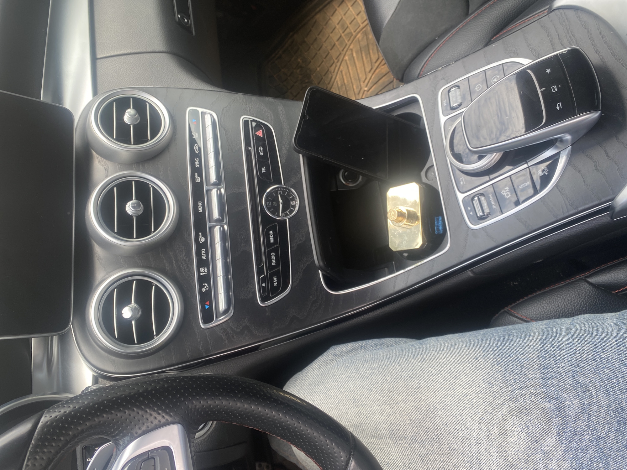 A close-up shot captures the interior of a car, likely a Mercedes-Benz, with a focus on the center console and dashboard. The steering wheel, upholstered in black leather with red stitching, is visible on the left. The center console features a wood-grain finish and houses a display screen, air vents, and various control buttons for climate, media, and navigation. A smartphone is placed in a storage compartment, and a golden rectangular object, possibly a fragrance atomizer, is also present. The gear shifter and surrounding controls are visible on the right. The background shows a glimpse of a textured floor mat and a black leather seat. The scene appears to be set in Oniyangi, Nigeria, based on the provided context.