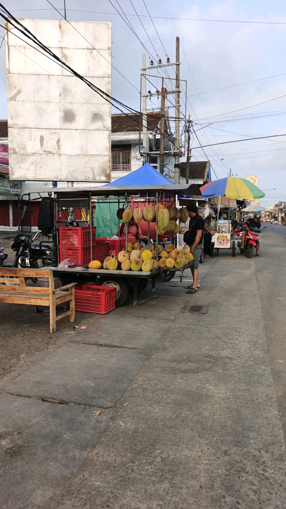The image shows a street scene in Suruhan Kidul, Indonesia, with a focus on a fruit stand. A truck bed is laden with durian fruits, some hanging and others displayed in piles. Several red plastic crates are visible, some empty and some containing fruit. A wooden bench sits to the left of the truck. A person in a dark t-shirt and shorts stands near the fruit display, possibly interacting with a customer. A woman in a pink shirt appears to be attending the stall as well. In the background, there are buildings with tiled roofs and utility poles with numerous wires. A motorcycle is parked to the left of the fruit stand, and another red motorcycle with a rider is further down the street. Two colorful umbrellas are set up over another vendor's cart in the distance. The ground is paved with concrete slabs. The overall setting suggests a typical roadside market or vendor setup.