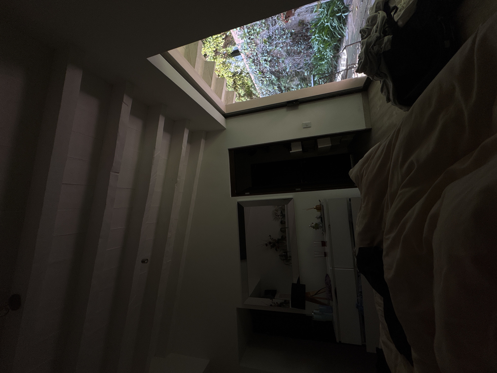 An interior shot depicts a dimly lit room looking upwards towards a large, rectangular open-air light well or skylight. The perspective suggests the viewer is at a lower level, gazing up into a vertical shaft.
On the left side, a prominent wall features architectural detailing with strong vertical elements, possibly columns or textured panels, creating depth and shadow. The right side of the room reveals a domestic setting: in the foreground, a large, light-colored blanket or bedding partially obscures the view, suggesting a bed or seating area. Beyond this, a wall section holds a mounted flat-screen television. Below the television, a rectangular mirror reflects a part of the interior, showing what appears to be decorative objects on a surface. To the right of the mirror, a tall, white cabinet or refrigerator is visible, with some items placed on its shelves.
The skylight or open-air shaft at the top of the image is brightly illuminated, showcasing lush green foliage and various plants, indicating an outdoor garden or courtyard above. On the left edge of this opening, concrete steps or structural tiers are visible, leading upwards.
The lighting within the room is subdued, with deep shadows, contrasting sharply with the bright, natural light streaming in from the overhead opening. This suggests either early morning, late evening, or an interior space designed to be dimly lit, relying primarily on the overhead light well for illumination. There are no people visible, and no discernible text. The scene conveys a sense of quietude and introspection in an urban dwelling in Mexico City, Mexico.
