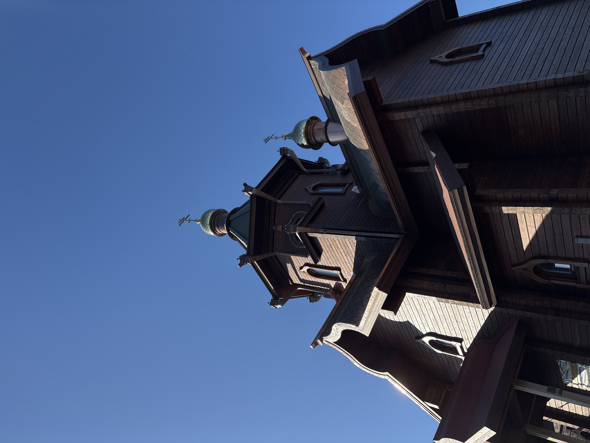 The image shows the wooden Pokrovsky Cathedral in Krasnoyarsk, Russia, viewed from a low angle, looking up towards the sky. The cathedral, constructed from dark wood, exhibits traditional Russian Orthodox architecture with multiple tiers and onion-shaped domes topped with crosses. The clear, bright blue sky contrasts with the building's dark color. The cathedral's wooden construction and details, such as the windows and roof structures, are visible. No people or other objects are present.