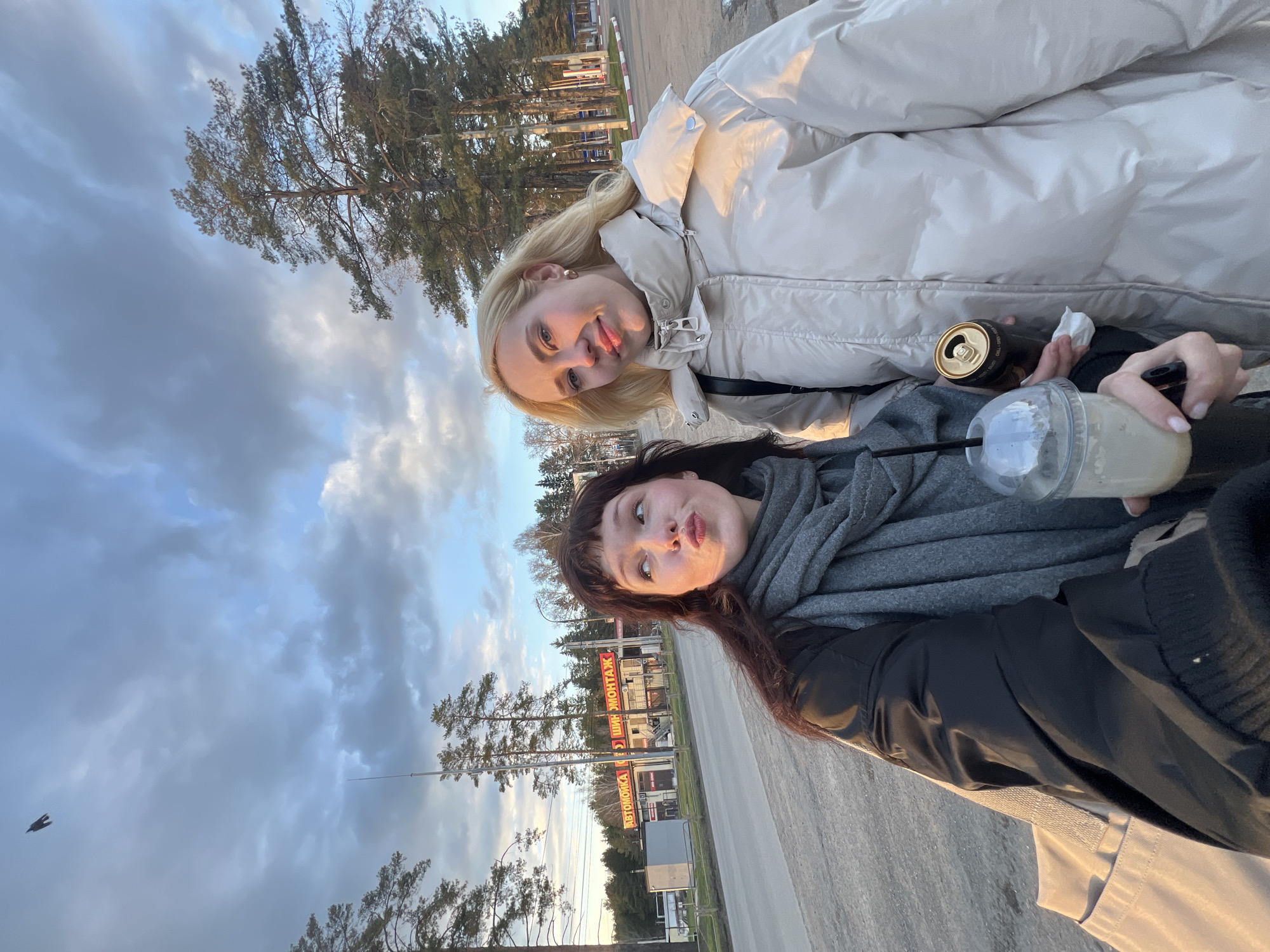 Two young women are captured in a selfie-style photograph outdoors, likely taken during the golden hour of late afternoon or early evening in Chaikovsky, Russia. The image is oriented with the horizon running vertically.
The woman in the foreground has dark hair, is wearing a grey scarf and a dark jacket, and is playfully pouting at the camera. She holds a clear plastic cup with a light-colored beverage and a straw. Beside her, a blonde woman in a light cream-colored puffer jacket smiles broadly. She holds an open black and gold can, possibly an energy drink, and a small white tissue.
The setting appears to be alongside a paved road or pathway, with tall pine trees lining the background. The warm, soft sunlight casts a glow on the subjects and the scene. The sky is partly cloudy with patches of blue visible. In the distance, several commercial buildings are visible with signs displaying Cyrillic text. Notable signs include "АВТОМОЙКА" (Car Wash) and "ШИНОМОНТАЖ" (Tire Fitting/Repair), indicating a roadside commercial area. A small bird is also visible flying in the sky. The overall mood is casual and friendly, suggesting a relaxed outing.