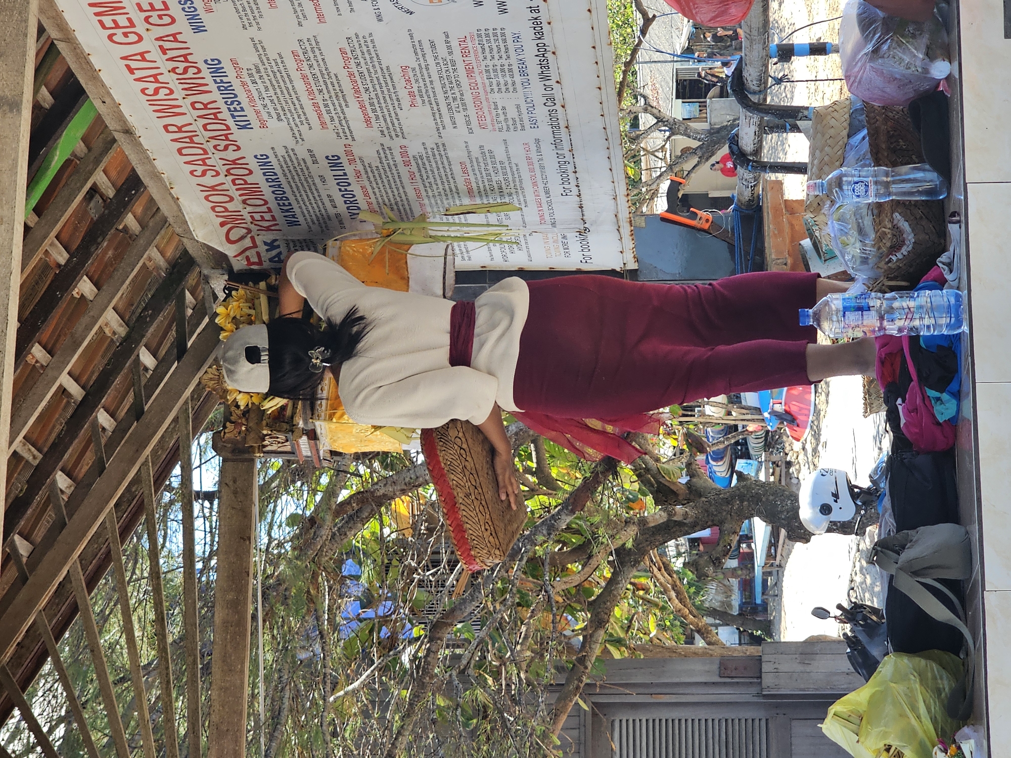 A woman wearing a white top, red sash, and red skirt stands facing a sign, holding a woven basket filled with offerings. She is positioned on a platform at a beachside establishment, likely in Denpasar, Indonesia, as indicated by the sign's text which includes "DENPASAR" and promotional information for watersports such as "KITESURFING," "WAKEBOARDING," and "HYDROFOILING." Behind the woman, a wooden canopy shelters the area, with tropical trees and vegetation visible. In the foreground, a collection of bags, a helmet, and water bottles are arranged on the platform. The overall scene suggests a service provider at a recreational beach location, with the woman possibly preparing to place the offerings as part of a ritual or tradition.