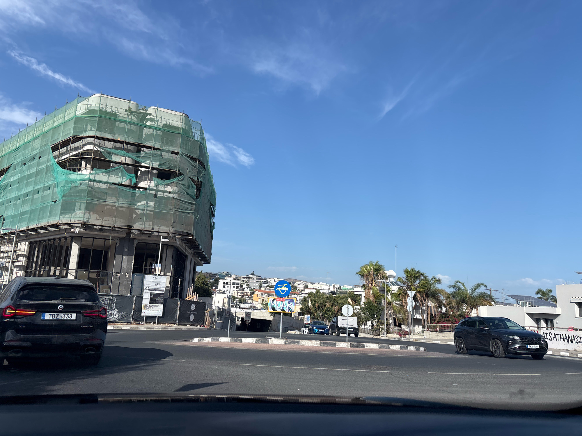 The image captures a street scene in Agios Athanasios Municipality, Cyprus. In the foreground, a black BMW SUV is parked, facing away from the camera. To the right, a dark-colored Hyundai SUV is in motion, turning. A building under construction, covered in green scaffolding and netting, dominates the left side of the frame. In the background, a cityscape with white buildings and palm trees is visible under a clear blue sky with wispy clouds. Traffic signs, including a blue circular sign with a white icon and a street sign with "MOA" graffiti, are present. The scene appears to be an urban environment during daylight.