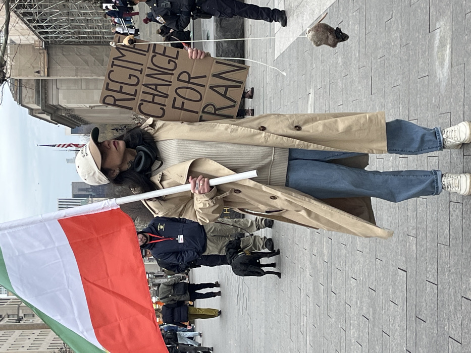 A long shot captures an outdoor public gathering in New York City, United States of America, specifically on a paved plaza in front of a grand neoclassical building with prominent steps, likely the Stephen A. Schwarzman Building of the New York Public Library on Fifth Avenue. The sky is overcast.
In the foreground, a person, appearing female, stands facing slightly right, holding a flag and a sign. The person wears a light-colored baseball cap, dark neckwear, a light beige ribbed sweater, a long tan trench coat, loose blue jeans, and white sneakers. Tattoos are visible on the left wrist and hand. Black headphones rest around the neck. In the left hand, a large flag on a white pole is held, displaying horizontal stripes of green, white, and red. In the right hand, a rectangular cardboard sign is held, inscribed with "REGIME CHANGE FOR IRAN" in dark lettering. A small, dark, furry object, resembling a dead rodent, hangs by a white string from the lower right corner of the sign, near the person's right leg.
In the mid-ground to the left, a man in a dark blue jacket and khaki pants stands with a black dog on a leash, possibly a K-9 unit. Other blurred individuals are visible walking. To the right, uniformed police officers stand near the steps of the background building. Numerous other individuals are distributed across the plaza and on the steps of the building. In the far background, an American flag flies from a pole, and multiple high-rise buildings pierce the skyline. The ground is paved with rectangular grey tiles.