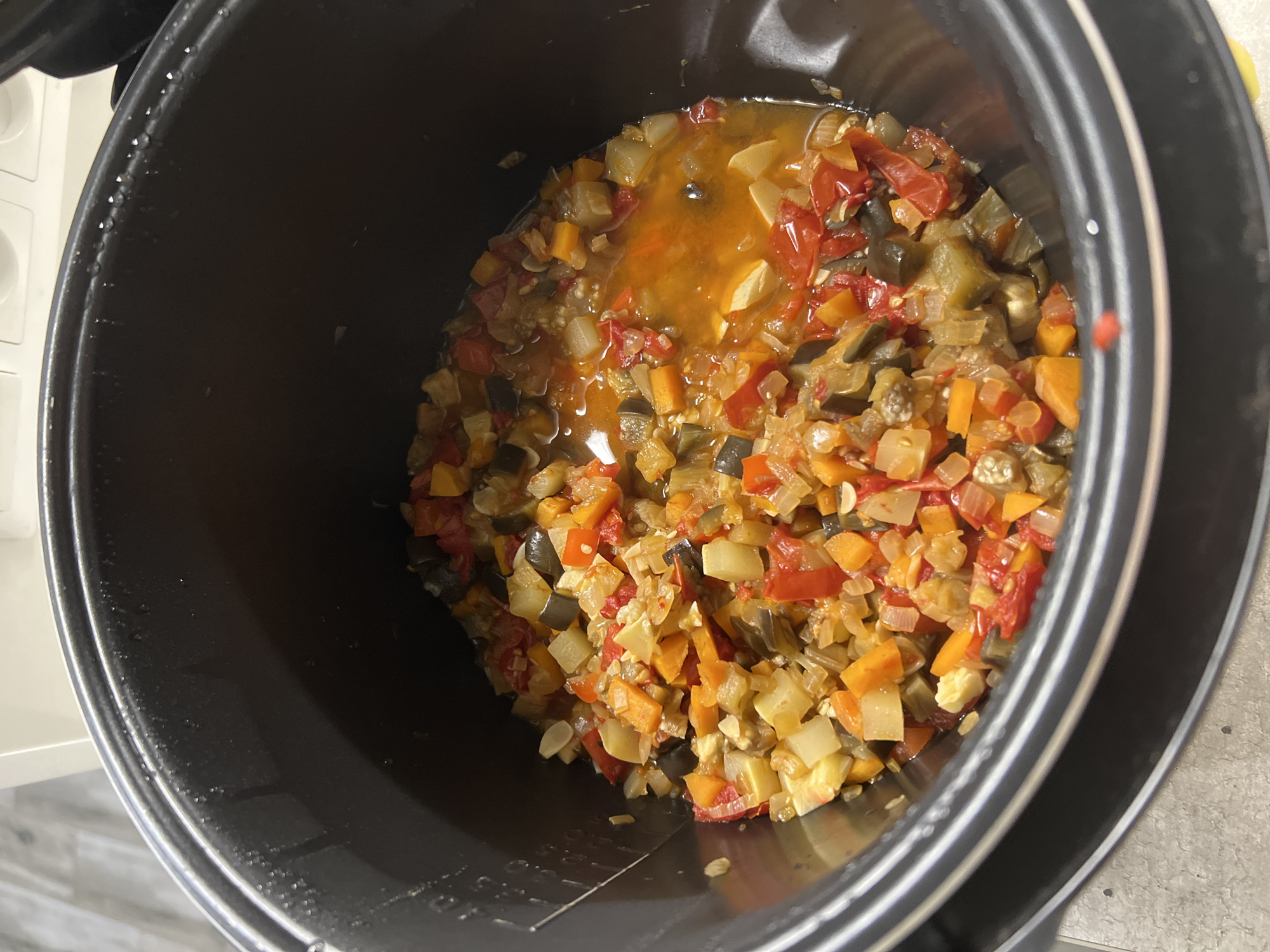 A close-up, top-down shot shows a black, electric multi-cooker pot filled with ratatouille. The stew is a colorful mix of diced vegetables including red bell peppers, eggplant, carrots, and what appear to be potatoes, all simmering in a reddish-orange broth. The interior of the pot shows water droplets on its upper rim. The surrounding countertop is a light gray speckled pattern. The image is a still-life, and no people or specific location in Volgograd, Russia are visible.