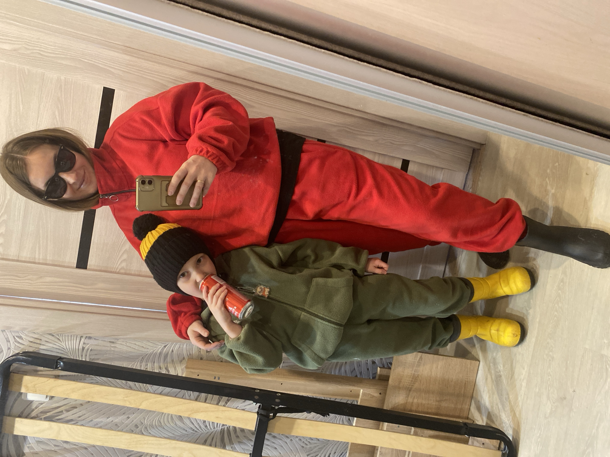 An indoor mirror selfie captured in Noviy, Russia, features an adult female and a young child. The adult female, positioned on the right and reflected in the mirror, has shoulder-length brown hair, wears black sunglasses, a red fleece zip-up top, red fleece pants, and a dark boot. She holds a gold-colored smartphone in her left hand, taking the photograph, and has her right arm around the child. A gold-colored ring is visible on her left ring finger. The child, a young boy, stands to her left, clad in a black knitted beanie with a yellow top and pom-pom, an olive-green fleece zip-up hoodie, matching pants, and bright yellow rain boots. He is observed drinking from a red can, which has a small charm attached. The background on the left displays a light-colored patterned wallpaper, against which a wooden bed frame or furniture component with black metal supports leans vertically. To the right, a light-colored wooden door with a black horizontal stripe is visible. The floor is light-colored laminate or wood.