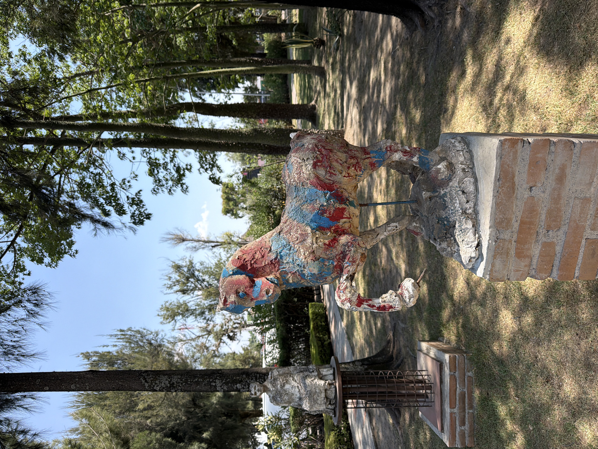 The image depicts a colorful, crudely painted concrete sculpture of a quadrupedal animal, possibly a dog or horse, standing on a stone base atop a brick pillar. The sculpture is painted with red, white, and blue pigments. The pillar is positioned in a grassy park area with mature trees casting shadows on the ground. In the background, there are more trees, and glimpses of buildings and structures can be seen. The location is most likely in the city of San Andrés Cholula, Mexico. There is also a second statue on the lower left of the image, this one is painted white.