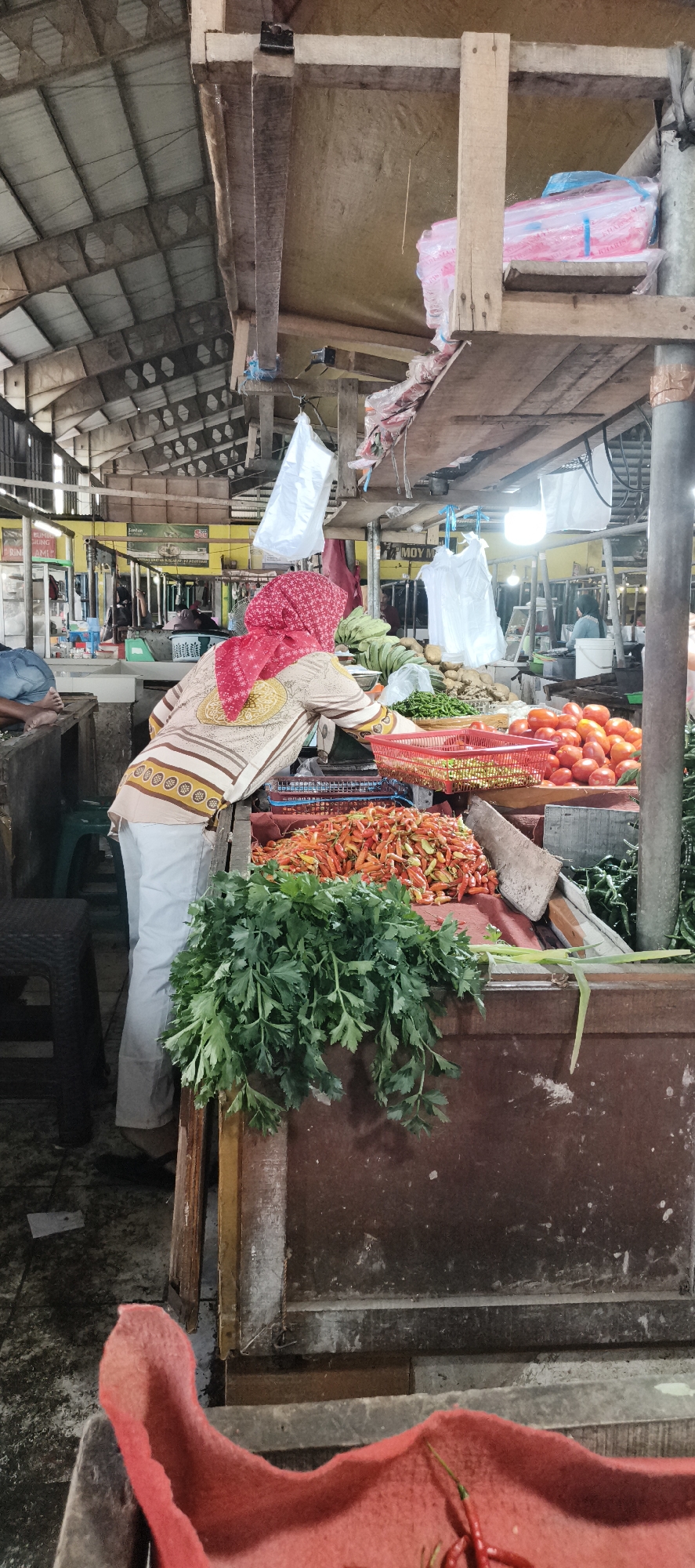 In Jambi, Indonesia, this image captures a vibrant scene within a bustling local market. A woman, wearing a red headscarf and a patterned tunic over light-colored pants, is seen from behind, leaning over a market stall laden with fresh produce. The stall displays an abundant variety of red chilies, tomatoes, and green vegetables, including bunches of fresh parsley. The background reveals the market's interior, characterized by a rustic wooden roof structure and hanging plastic bags. Other market-goers are visible in the distance, and various goods are displayed on shelves and stalls. The overall atmosphere suggests an active and authentic Indonesian market environment.