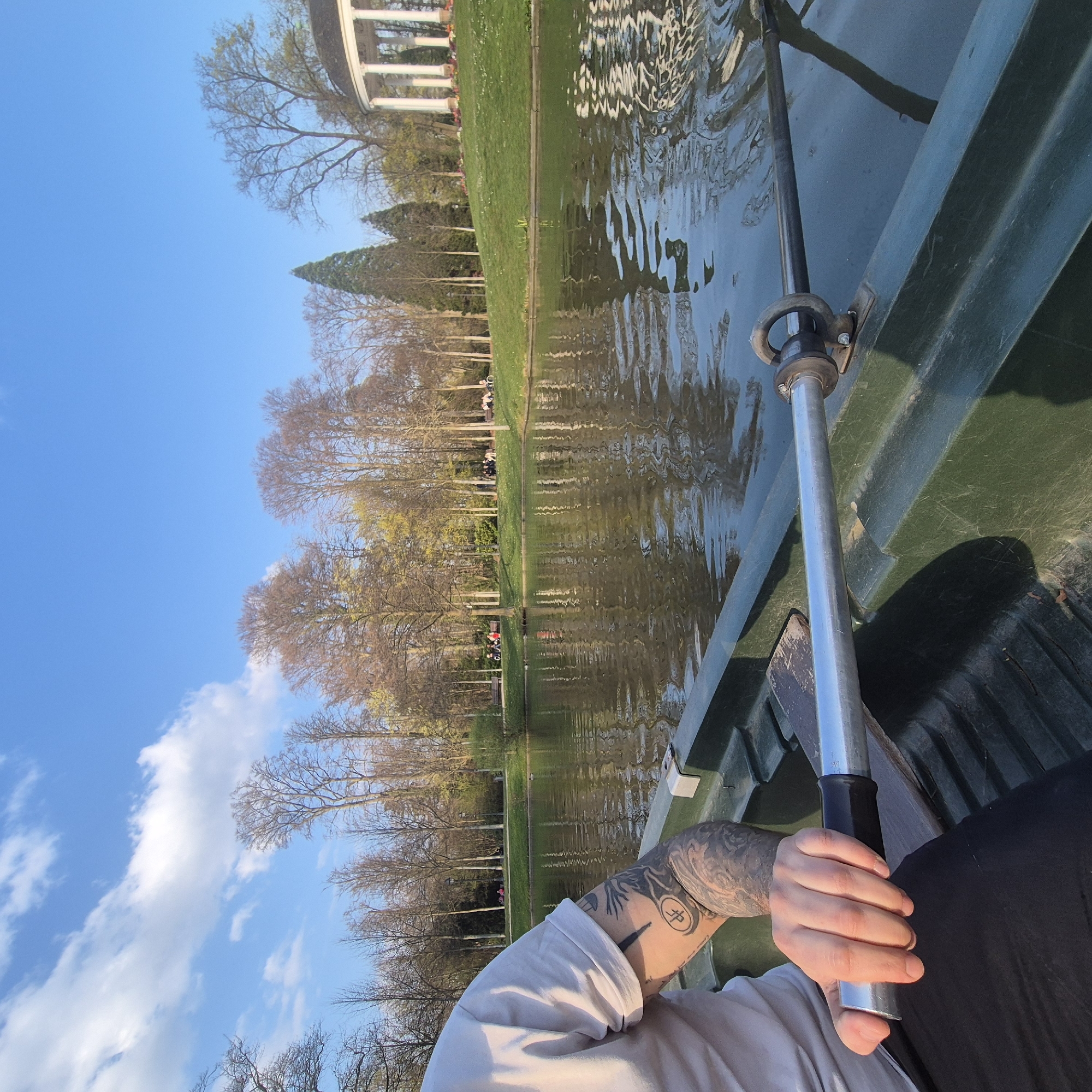 The image presents a first-person perspective from inside a dark green rowboat on the lake within the Parc de l'Orangerie in Strasbourg, France. In the lower left quadrant, the torso and extensively tattooed left arm of an individual are visible, clad in a light-colored, short-sleeved t-shirt. The arm displays a full sleeve tattoo and a distinct circular symbol on the inner bicep/forearm. The person's hand firmly grips the silver metal shaft of an oar, which features a black rubber grip, the oar secured in an oarlock on the boat's gunwale.
The calm surface of the lake occupies the midground, reflecting the sky and the surrounding parkland with subtle ripples. Across the water, a gently sloping grassy bank leads to a paved pathway. This pathway is lined with numerous deciduous trees, many still bare or just beginning to bud, indicative of early spring foliage. Several individuals and small groups are visible on the pathway and grassy areas, some seated on benches. To the far right, situated on an elevated grassy mound, a distinctive white classical rotunda with columns, identified as the Temple de l'Amour, stands prominently. The sky above is a clear blue with scattered white cumulus clouds.