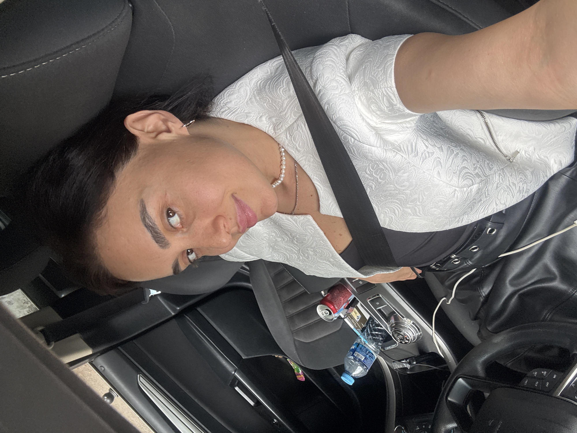 In Tbilisi, Georgia, a woman is seated in the driver's seat of a car. She is looking towards the camera, taking a selfie. She is wearing a white textured blazer over a black top, a pearl necklace, and small hoop earrings. A black leather skirt with a silver-buckled belt is visible. Her seatbelt is fastened across her chest. In the car's center console area, a bottle of water, a can of soda, and cigarette packs are present. The car's interior, including the steering wheel and gear shift, is visible.