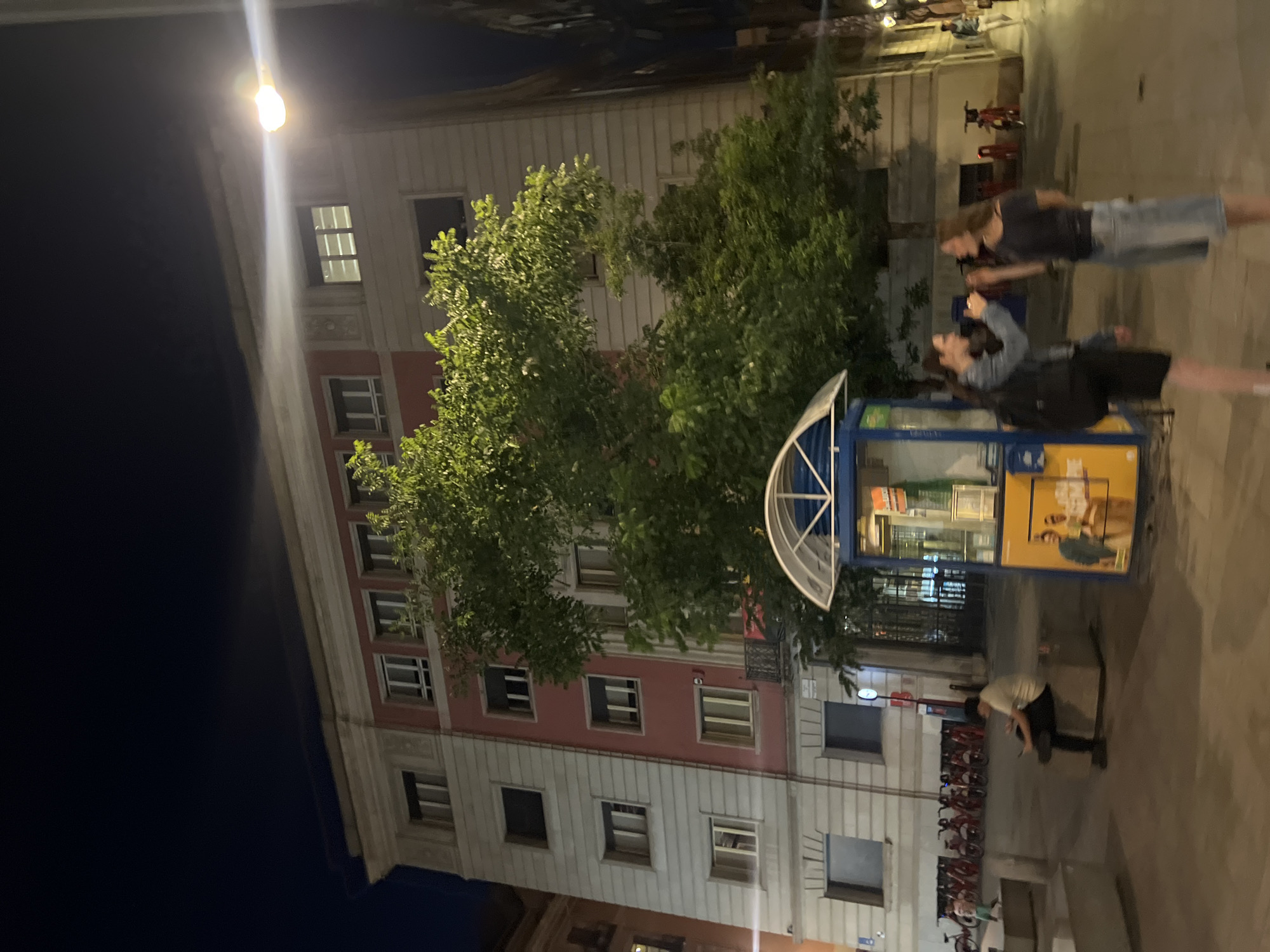 The image shows an outdoor scene at night on a city street in Barcelona, Spain. In the foreground, two young women are standing and talking, with one holding a drink. Behind them is a blue and yellow kiosk with an arched canopy. To the left of the kiosk, several red rental bicycles are parked in a row. Further to the left, a person is sitting on a concrete block, looking at their phone. In the background, a large building with a pink and white facade, featuring multiple windows, stands tall. Trees with green foliage are in front of the building. A streetlamp with a bright light is visible on the right side of the image, illuminating the scene. The ground is paved.