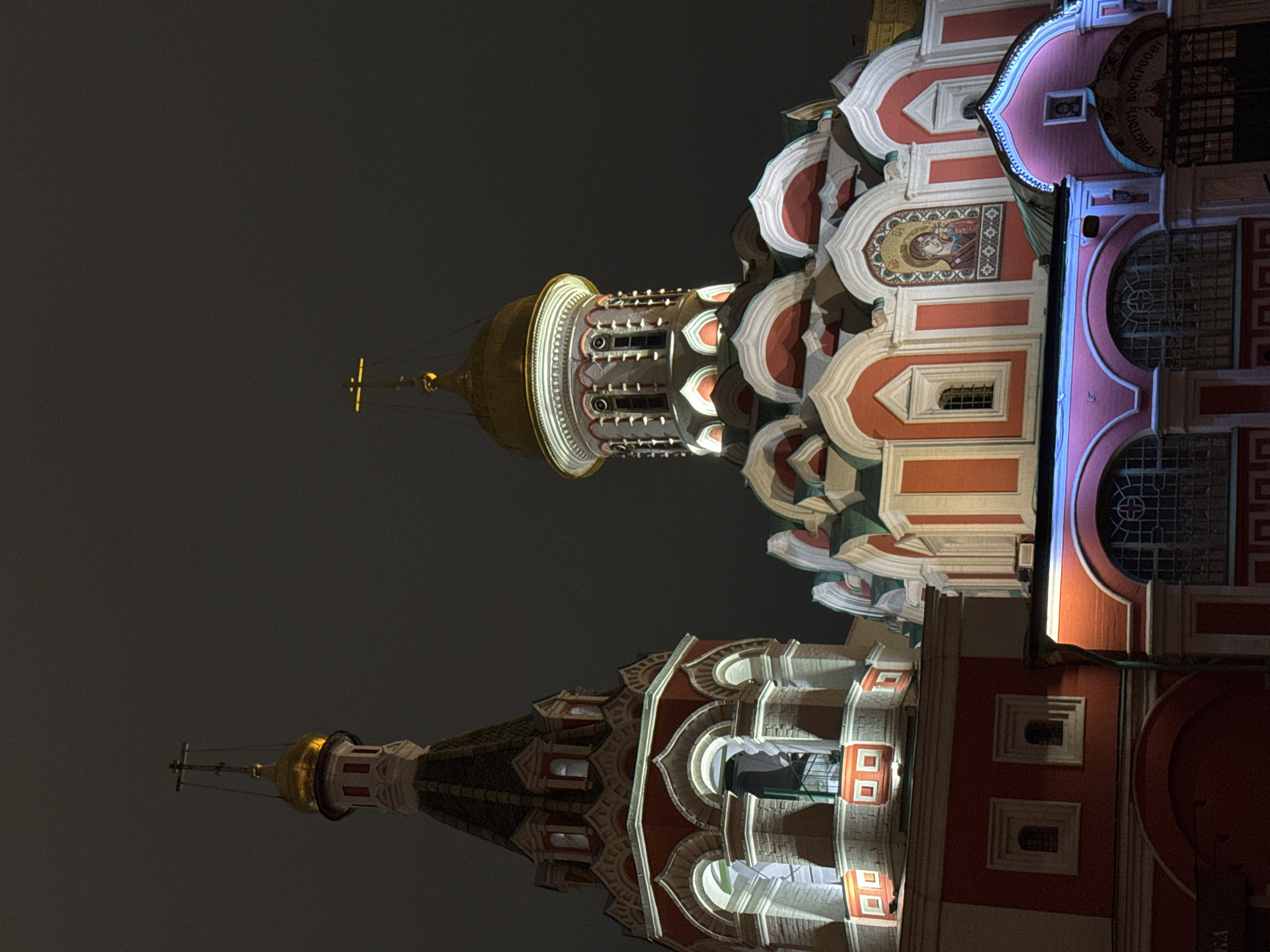 The image shows a nighttime view of the architectural details of a building in an unidentified city. The architecture features elements such as a tower with a golden dome, crosses, and decorative ornamentation in shades of red, white, and gold. The building is illuminated with lights that highlight the architectural details. The sky is dark and featureless.