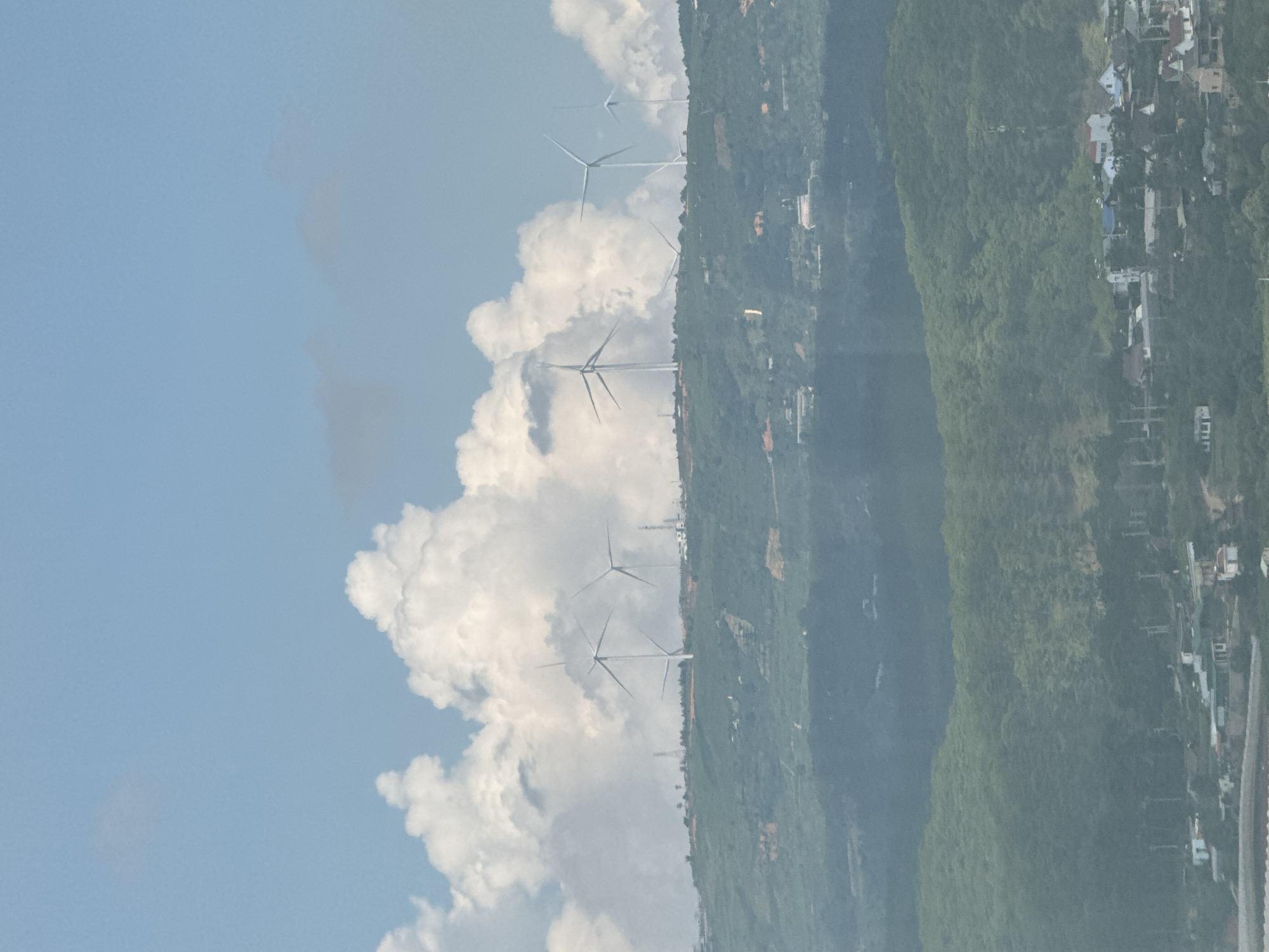 The image depicts a landscape with a clear blue sky and scattered white clouds, above rolling green hills. Several wind turbines are visible on the crest of the hills, with their blades spread. In the foreground and further down the hills, buildings and houses are scattered amongst the greenery, suggesting a settlement. The overall scene appears to be a rural or semi-rural area with evidence of wind energy infrastructure. The location is Đa Lộc, Vietnam.