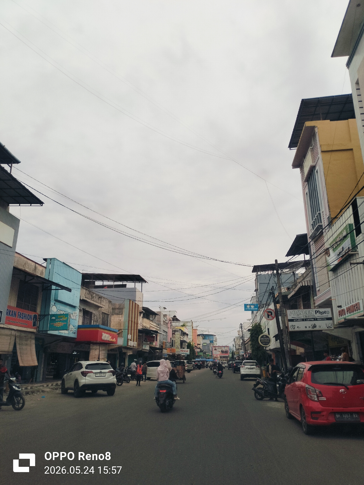 The image depicts a street in Lhokseumawe, Indonesia, on an overcast day. The street is lined with two-story buildings, many of which have shops on the ground floor. Signage is visible in Bahasa Indonesian. There are cars and motorcycles driving on the road and parked along the sides. Power lines crisscross above the street. Pedestrians are also visible. The camera information indicates the image was taken with an OPPO Reno8 device.