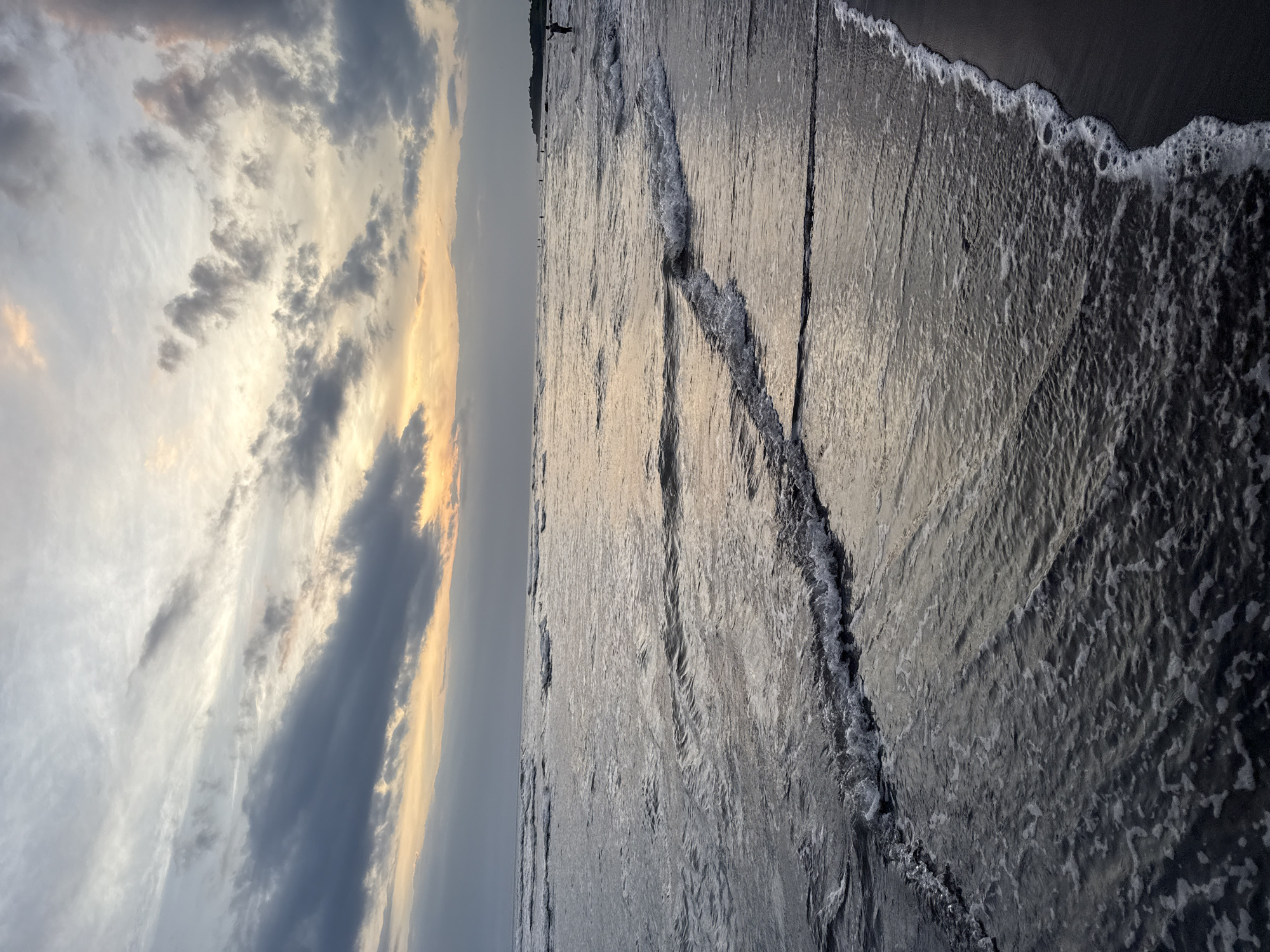 The image shows a beach at sunset, possibly in Unknown, Unknown. The sky is filled with clouds, some dark and some illuminated by the setting sun. The sun's light reflects on the water, creating a shimmering effect. Waves are visible, approaching the shore, with foamy edges. The sand is dark and wet. A person stands far in the distance on the beach.