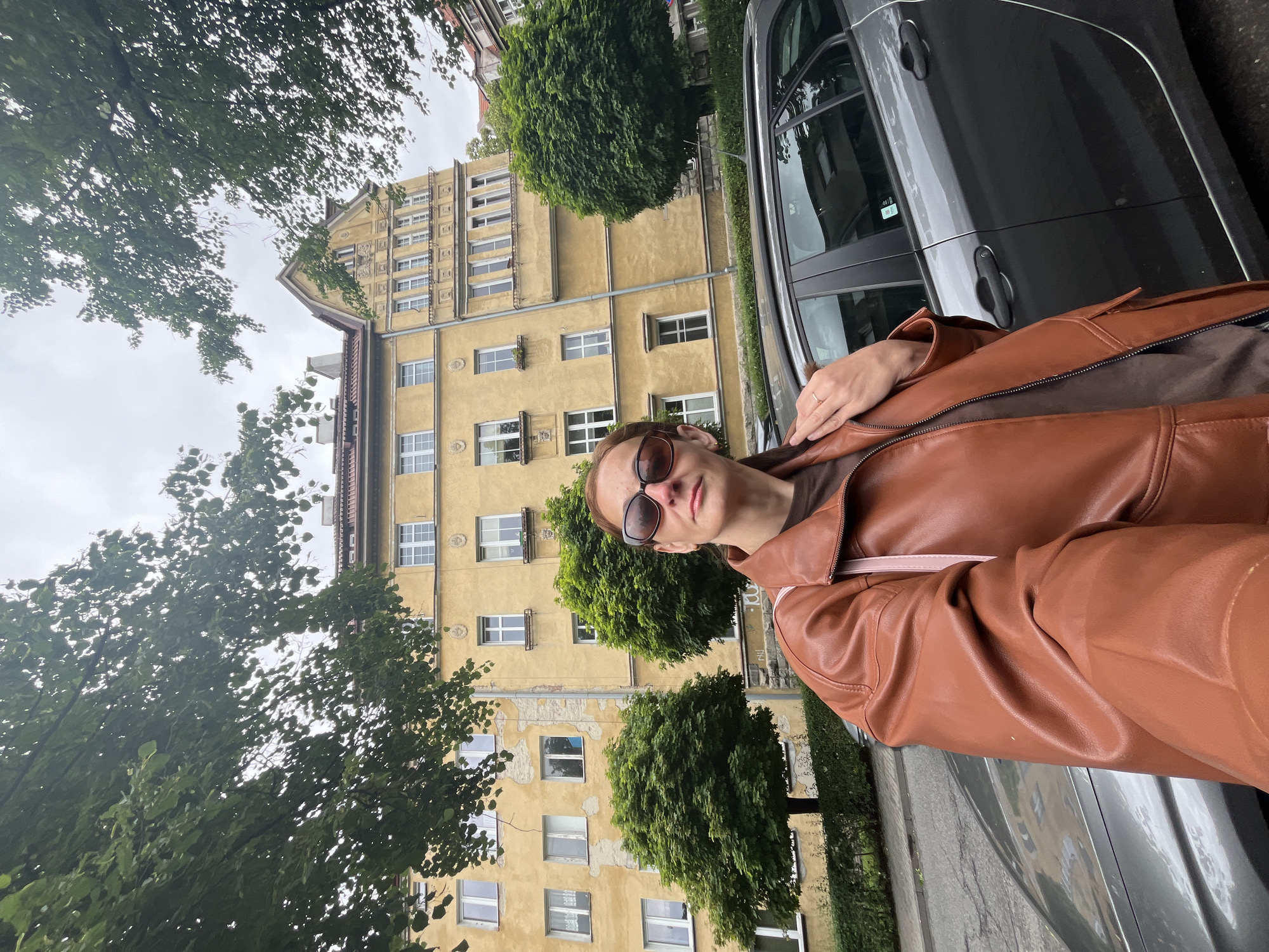 A woman stands in front of a yellow apartment building with trees and a car. She is wearing a brown leather jacket, sunglasses, and a brown shirt. The building has many windows and decorative architectural details. The car is dark gray. The sky is overcast.