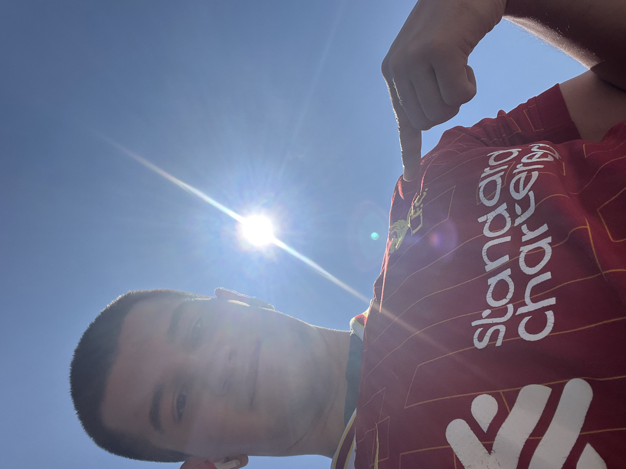 A low-angle, outdoor selfie captures a person wearing a red soccer jersey with white lettering "Standard Chartered" on the front. The person is pointing to the logo on the jersey. The background shows a bright blue sky with the sun shining brightly, creating lens flare. The person appears to have short, dark hair. The location and the details of the city of Varna, Bulgaria, are not apparent.