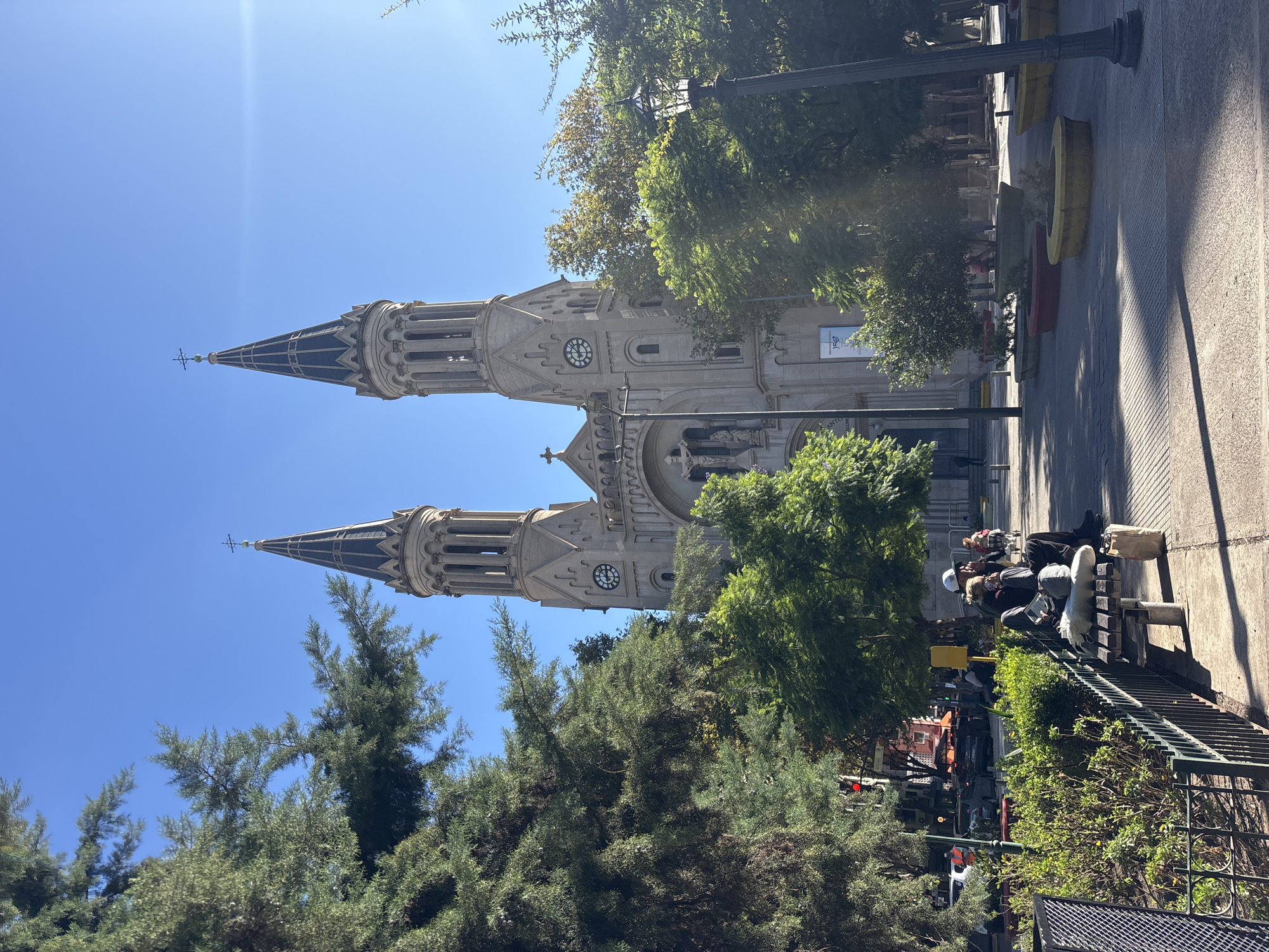 A daytime photograph, rotated 90 degrees clockwise, captures a grand church with two prominent spires dominating an urban scene, likely a public plaza or park in Buenos Aires, Argentina. The impressive church, possibly of neo-Gothic architecture, features two tall, ornate spires, each topped with a cross and displaying a clock face. Its facade includes detailed stonework, arched windows, and statues, with a prominent white banner hanging on the lower part of the right tower. The banner clearly reads "150 AÑOS" (150 Years), and below it, partially visible text includes "SANTO" and the date "1880," suggesting a significant anniversary.
The foreground and midground are densely populated with green trees, some partially obscuring the church. To the right, a wide paved area, possibly a pedestrian walk or plaza, is lined with wooden benches. Several people are seated on these benches, appearing to relax or socialize in the sunshine. A metal fence separates the seating area from a street in the background, where a red traffic light and a few vehicles are visible.
The scene is bathed in bright sunlight under a clear blue sky, casting distinct shadows from the trees, lampposts, and people, indicating late morning or early afternoon. Decorative, colorful concrete planters (yellow and red) are spaced along the paved area. The overall impression is one of a peaceful, bustling public space, combining historical architecture with everyday urban life.