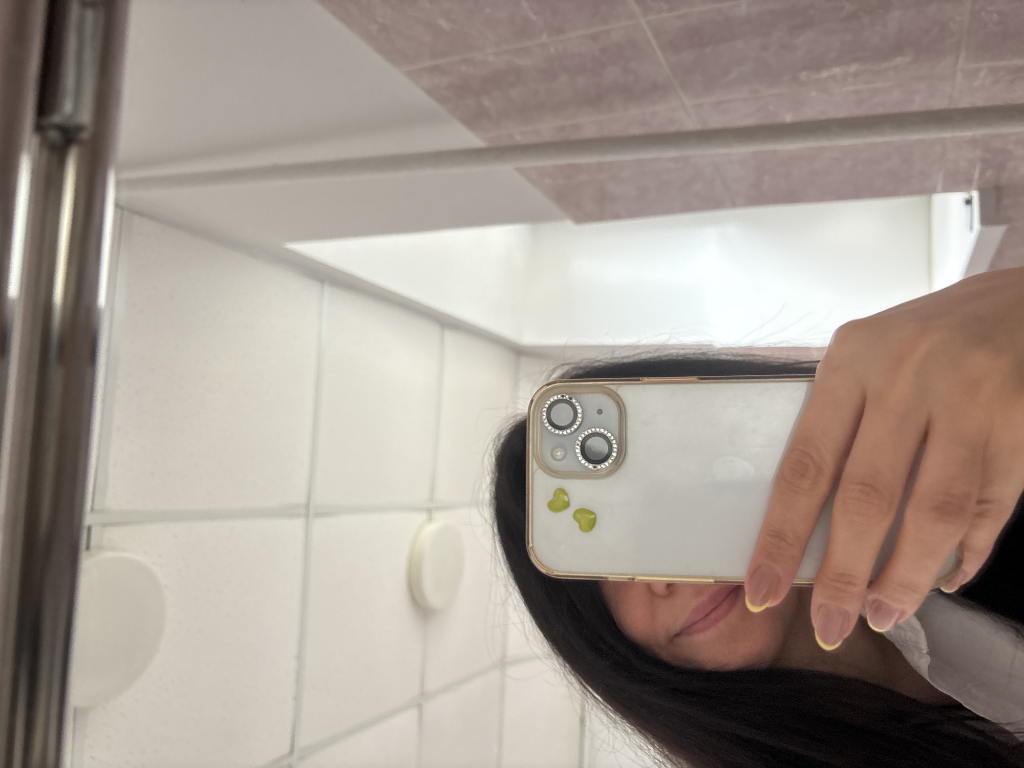 A person is taking a selfie in a bathroom mirror. The bathroom features white tile walls and a pink tile ceiling. The person is holding a white phone with a gold-colored case, embellished with decorative hearts near the camera lens. Part of the person's face, dark hair, and hand are visible. The location of the city of Chaikovsky, Russia is not indicated.