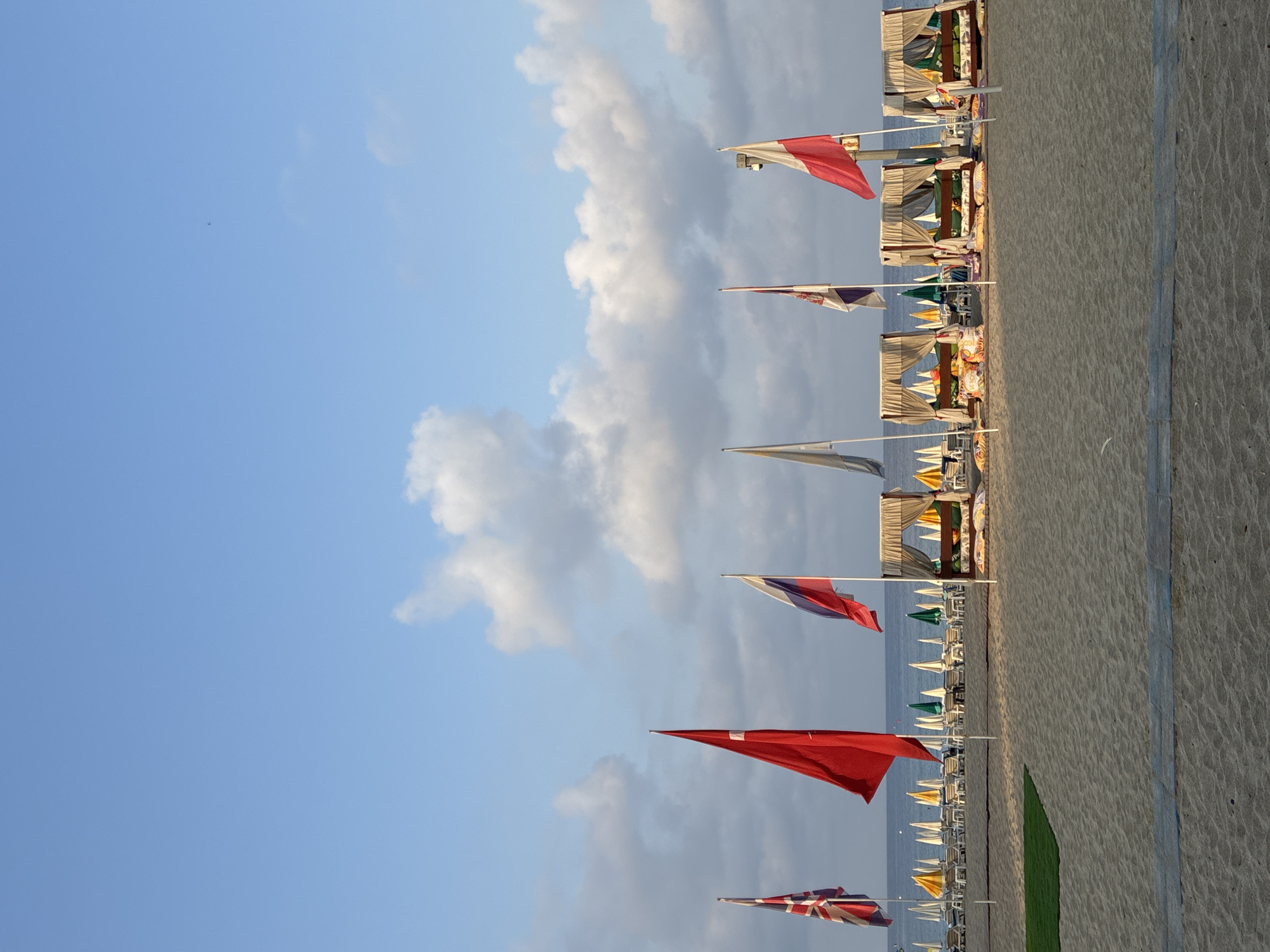 An image captured from a sandy beach in Alanya, Turkey, shows a row of flags flying against a sky filled with scattered clouds. From left to right, the visible flags include the Union Jack, the Turkish flag, the Russian flag, and the Polish flag. Behind the flags, a line of beach chairs and umbrellas stretches along the shoreline, with the calm blue sea visible in the distance. In the foreground, several draped cabanas with cushions are set up on the sand, suggesting a resort or leisure area. The overall scene depicts a peaceful beach setting.