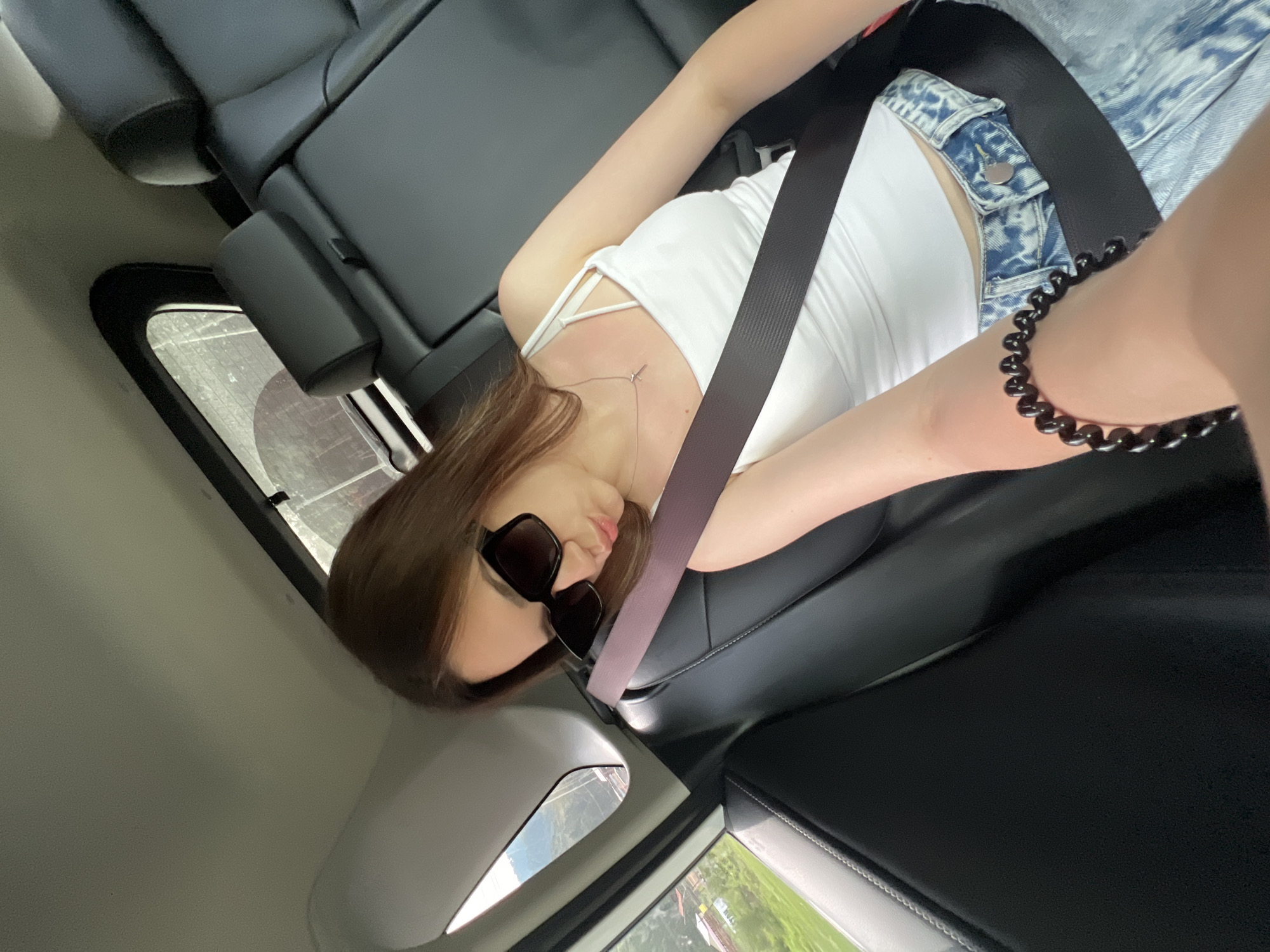 A young woman is in the back seat of a car, wearing a white tank top, blue jeans, sunglasses, and a cross necklace. Her left arm is extended towards the camera, with a black spiral bracelet on her wrist. She is leaning her head on her arm, with her lips pursed. The seatbelt is across her chest. The car's interior, including black leather seats and a window, is visible. Outside the window, a green landscape with some buildings can be seen. The image appears to be a selfie taken inside the moving vehicle.