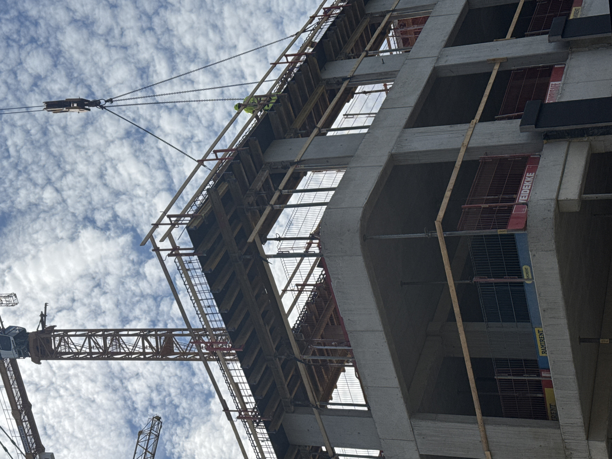 The image captures a construction site in Tallinn, Estonia, on a cloudy day. A large tower crane dominates the upper left of the frame, with its jib extending horizontally and a prominent boom extending vertically. Below the crane, a multi-story concrete building is under construction, featuring unfinished floors, exposed rebar, and scaffolding. A worker in a high-visibility yellow jacket and helmet stands on a temporary wooden platform on one of the upper levels, near a section of the crane's lifting apparatus. Metal safety barriers, some red and some blue, are visible along the edges of the unfinished floors. Signs for "RAMIRENT" and "EIDEKKE" are present on some of the barriers, indicating equipment rental and construction company services. The overall scene depicts ongoing construction work with heavy machinery and workers actively involved in the building process.