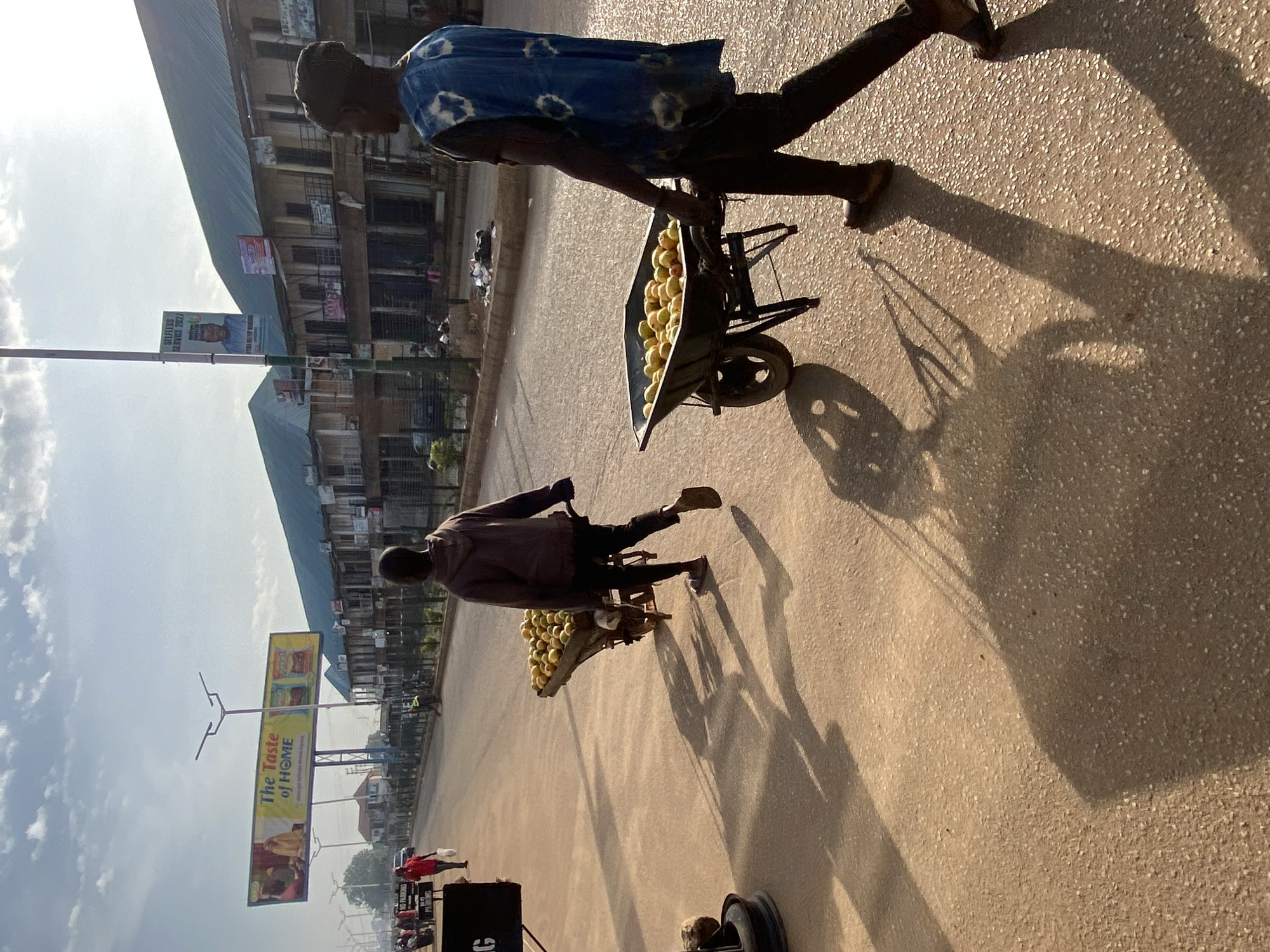 In Bukuru, Nigeria, two individuals are seen pushing wheelbarrows loaded with yellow fruits along a sunlit street. The person in the foreground, wearing a blue and white tie-dye vest and dark trousers, pushes a wheelbarrow filled with what appear to be mangoes or oranges. Behind them, another individual, dressed in a dark hoodie and trousers, pushes a similar wheelbarrow also laden with fruits. The street is paved and appears to be a commercial area, with buildings lining the right side of the road, featuring balconies and shopfronts. Several billboards are visible in the distance, including one advertising "The Taste of HOME". The sunlight casts long shadows across the street, indicating it is either early morning or late afternoon. There are a few other people and vehicles visible further down the street, suggesting ongoing activity in the area.