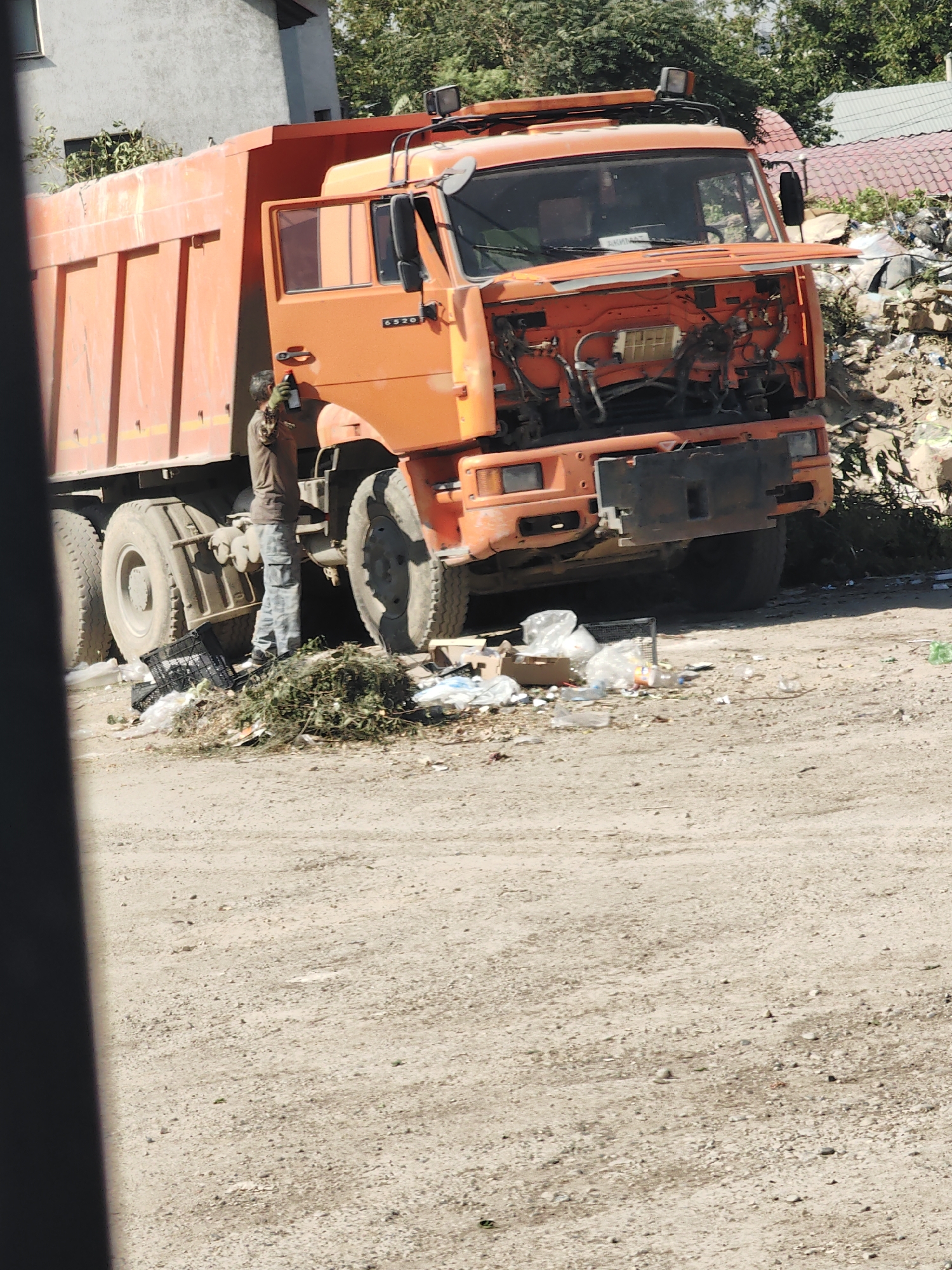 An orange dump truck is parked on a dirt surface, its hood open. A person wearing a brown shirt and gray pants is standing next to the truck, holding a spray bottle and seemingly attending to it. The area around the truck is littered with debris, including plastic bags, bottles, and vegetation. In the background, there is a building and trees. The scene appears to be an outdoor, possibly industrial or construction, location.