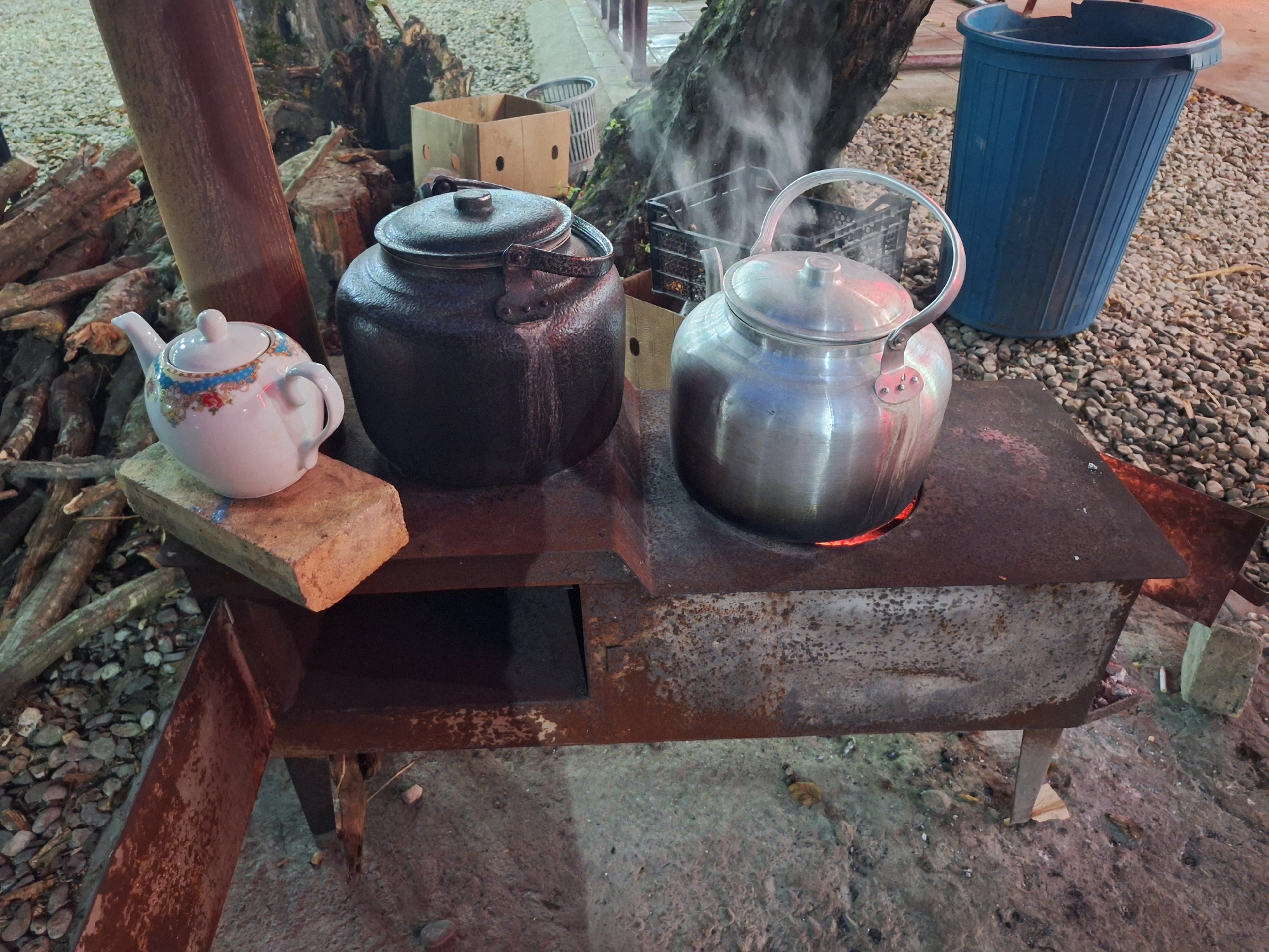 The image depicts an outdoor scene in Nabran, Azerbaijan, featuring a rustic, metal cooking stove. Two large kettles are actively heating on the stove, with steam rising from the one on the right. To the left, a smaller, decorative white teapot rests on a brick. In the background, a pile of firewood is visible, along with cardboard boxes and a blue trash bin. The ground is covered with gravel and dirt. The scene conveys a sense of outdoor cooking or preparation, likely in a rural or less developed setting within Nabran.