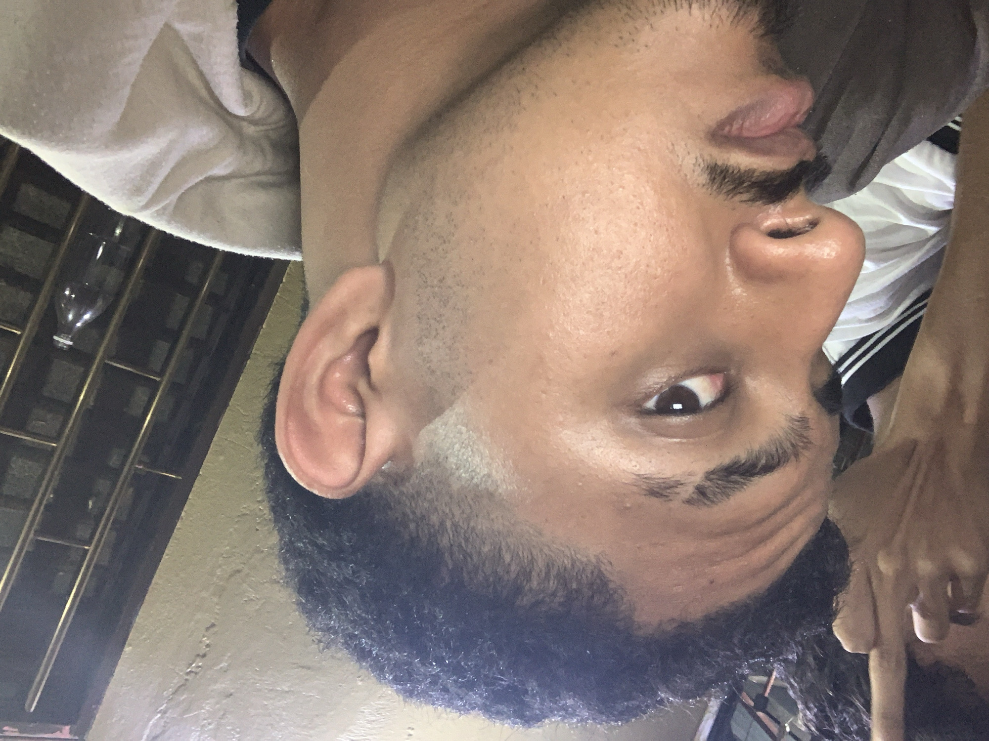 The image is a close-up, side-profile shot of a young man, appearing to be in his late teens or early twenties. He has dark, curly hair styled in a fade haircut. His skin is light brown, with a visible light mustache and a hint of stubble on his chin. His left eye is visible, brown in color, and he appears to be looking towards the camera. His left ear is also visible.
He is wearing a dark t-shirt and what appears to be a light-colored hooded jacket or sweatshirt. In the background, another person's arm and hand are visible, with the hand making an offensive gesture. The background also shows a wall and a barred window with a plastic bottle visible through it. The lighting suggests it is nighttime or indoors with artificial lighting.
The image is identified as being taken in Santa Ana, Venezuela.