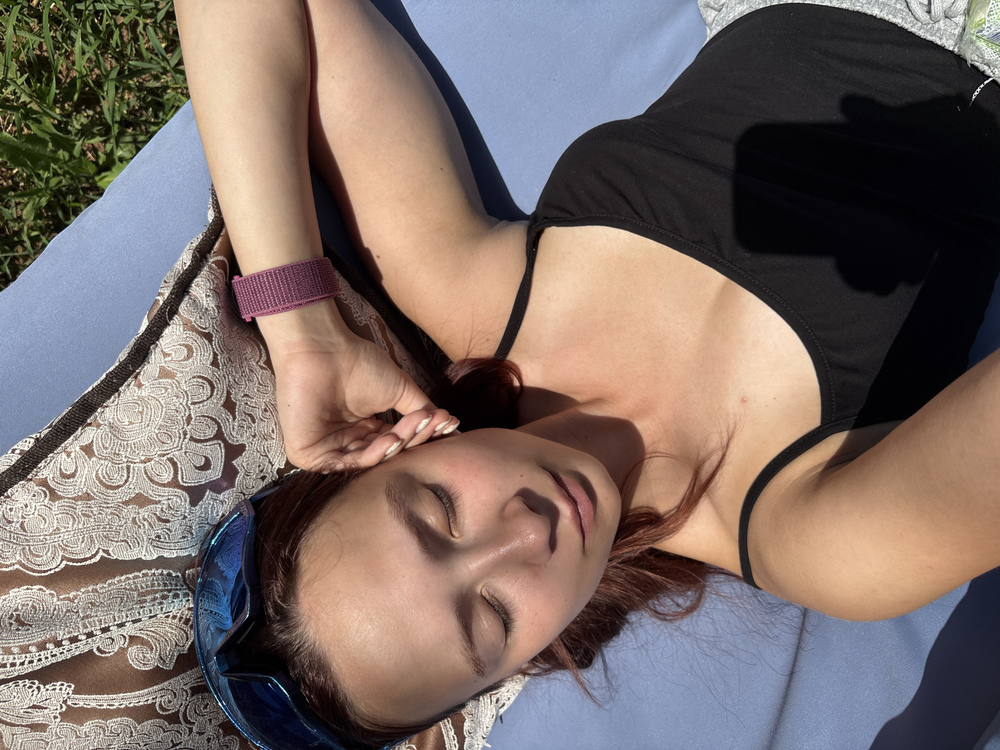 A young woman with long, dark red hair is lying on her back on a blue mat outdoors. She is wearing a black tank top and a purple bracelet on her left wrist. Blue ski goggles are positioned near her head on the mat. Her eyes are closed, and her head is tilted slightly to her right, with her right hand resting on her forehead. The mat is placed on the ground, with green grass visible in the background. The scene appears to be in a sunny outdoor environment, possibly a park or backyard, given the presence of grass and natural light. There is no visible indication of the specific location within Прикамский, Russia.
