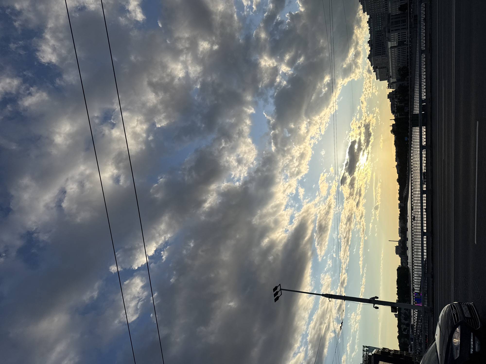 This is a vertical, medium-shot, outdoor photograph taken from a moving vehicle, capturing a city skyline and a dramatic sky at dusk. The foreground shows the edge of an asphalt road and the hood of a black car. Beyond the road, there is a bridge with a decorative railing. A river or body of water runs behind the bridge, lined with trees and buildings. A tall street lamp with three lights is visible on the left side of the frame. The sky is filled with a mix of dark and light clouds, with the setting sun peeking through, casting a warm glow. Several dark, horizontal power lines stretch across the top of the image. The overall impression is one of urban transition at the end of the day.