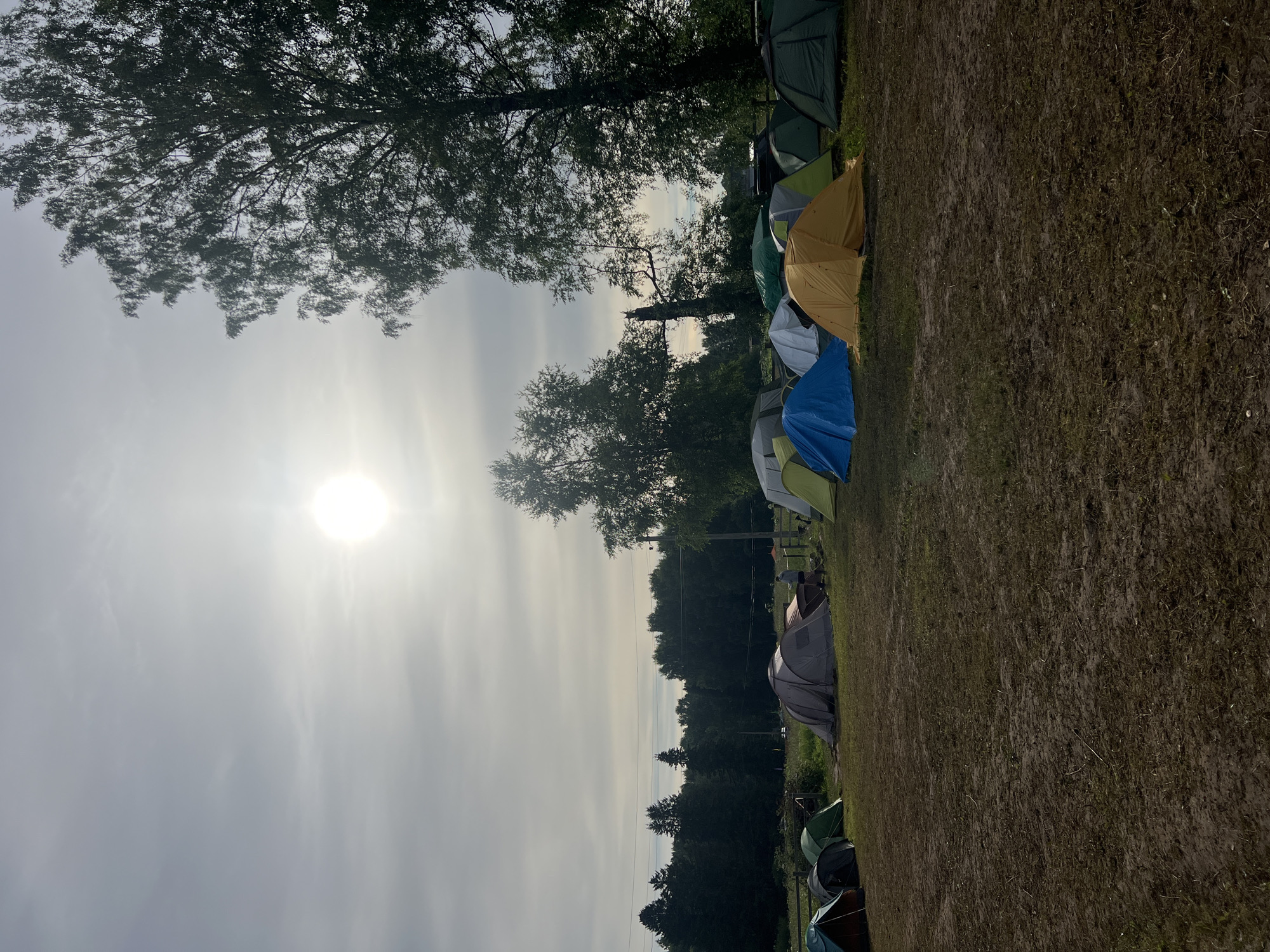 This media file depicts an outdoor scene with a campsite in Сергино, Russia. The primary subjects are a row of tents set up on a grassy, somewhat muddy field. The tents vary in color, including green, yellow, blue, and grey. In the background, there is a dense line of trees forming a forest, with more trees silhouetted against a hazy sky.
The time of day appears to be morning or late afternoon, given the bright but diffused sunlight filtering through the clouds and casting a soft glow on the scene. The weather seems overcast or hazy, contributing to the muted light.
No specific activity or event is clearly depicted, but the presence of multiple tents suggests a camping event or a festival. There is a sense of quiet and stillness in the image, with no visible people. The ground appears uneven and damp in places, indicating recent rain or morning dew.
There is no visible text in the image. The overall mood is tranquil and natural, focusing on the camping setup amidst a serene, natural landscape.