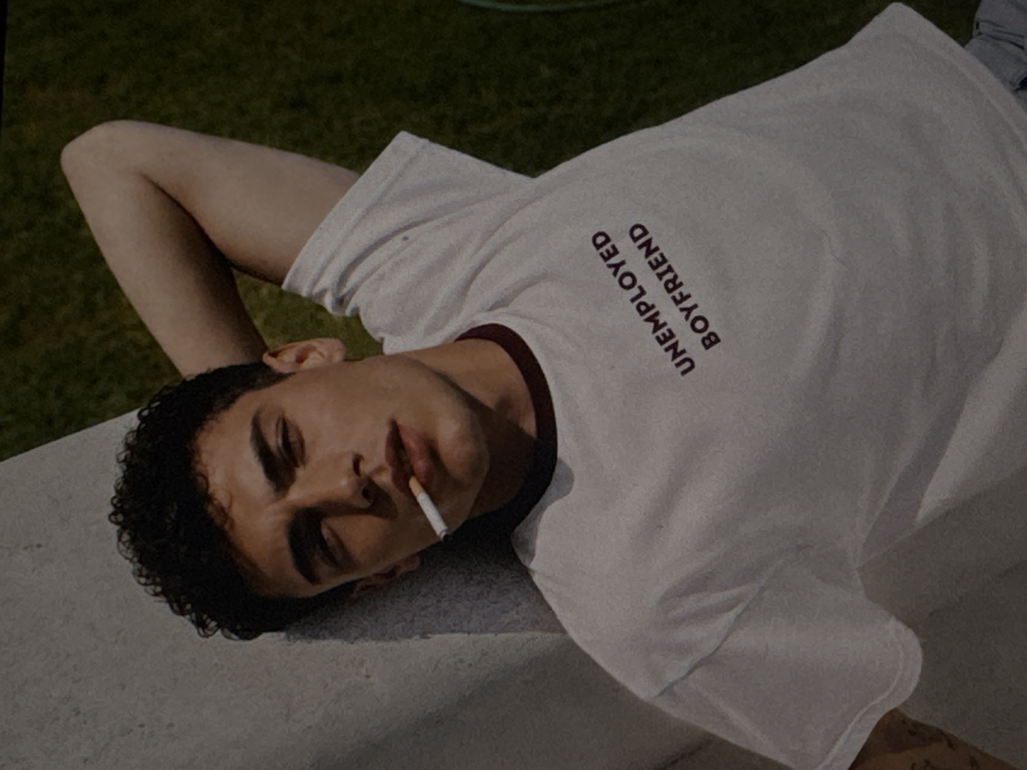 The media file shows a close-up, angled shot of a young person lying down on a textured surface, possibly a wall or a bench. They are wearing a white t-shirt with maroon trim around the collar. Printed on the t-shirt in a dark, bold font, oriented vertically and reading from bottom to top, is the text "UNEMPLOYED BOYFRIEND". The person has dark, curly hair and is holding a cigarette in their mouth, which appears to be lit. Their eyes are closed or partially closed, and their facial expression is relaxed, possibly conveying a sense of weariness or contemplation. The background is out of focus, appearing as a blurry green area, suggesting an outdoor setting with vegetation. The lighting suggests it is daytime, with shadows indicating the sun's presence. No specific landmarks are visible to pinpoint the location within Kuta Selatan, Indonesia, though the outdoor setting is consistent with tropical environments.