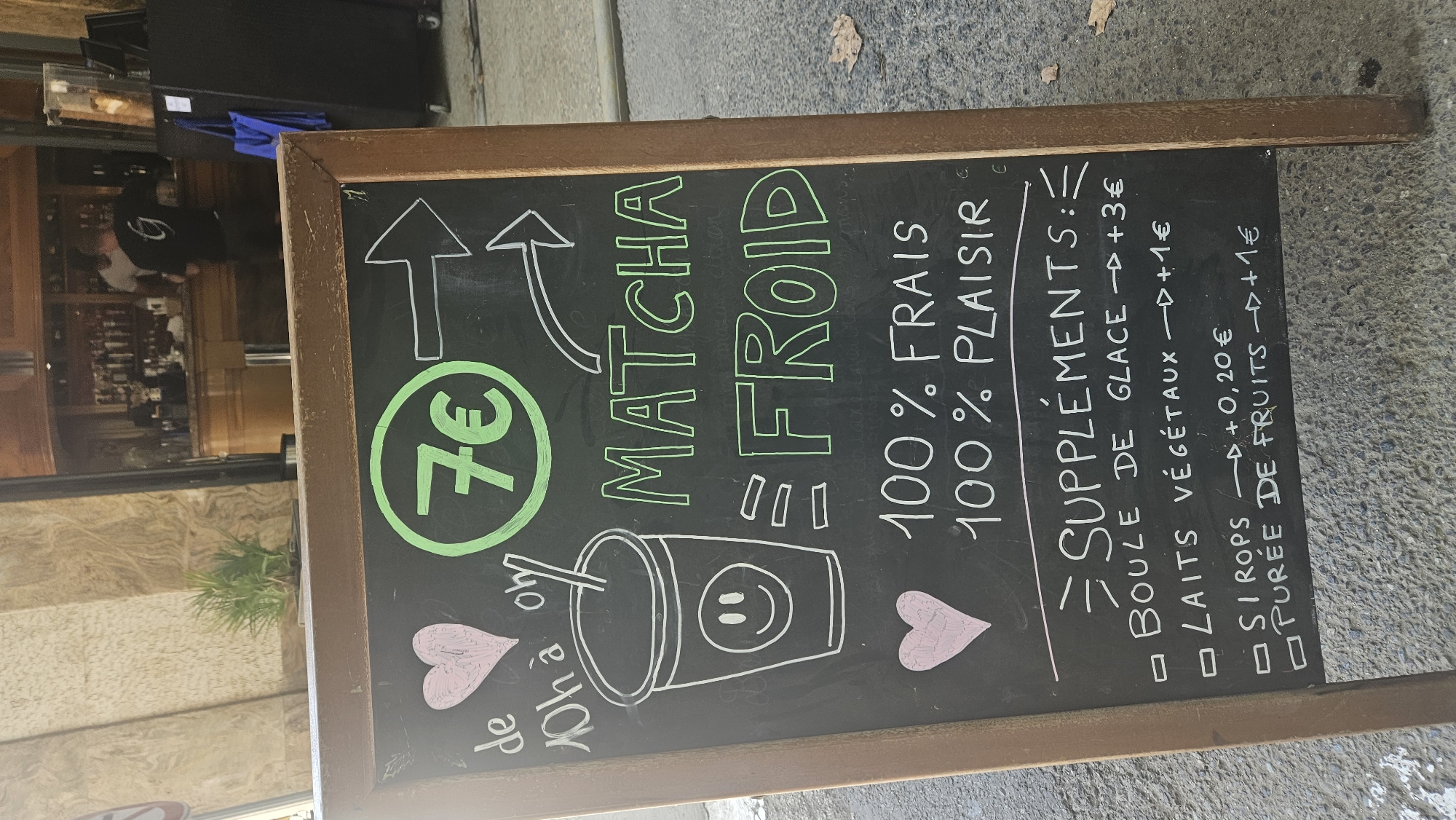A close-up shot captures a chalkboard sign in L'Île-Rousse, France, advertising a cold matcha drink. The sign is framed by a rustic wooden border and rests on a textured gray surface, likely pavement. At the top, the price of "7€" is prominently displayed in a green circle. Below this, the words "MATCHA FROID" are written in bold, green lettering. Further down, the sign promises "100% FRAIS" (100% fresh) and "100% PLAISIR" (100% pleasure), with two pink heart drawings adding a touch of visual appeal. A section titled "SUPPLÉMENTS" (Supplements) lists options such as "BOULE DE GLACE" (scoop of ice cream) for an additional 3€, "LAITS VÉGÉTAUX" (plant-based milks) for 1€, "SIROPS" (syrups) for +0,20€, and "PURÉE DE FRUITS" (fruit purée) for +1€. The background is slightly blurred, showing a glimpse of a building facade with cream-colored stone and hints of an indoor establishment with shelves and bottles.