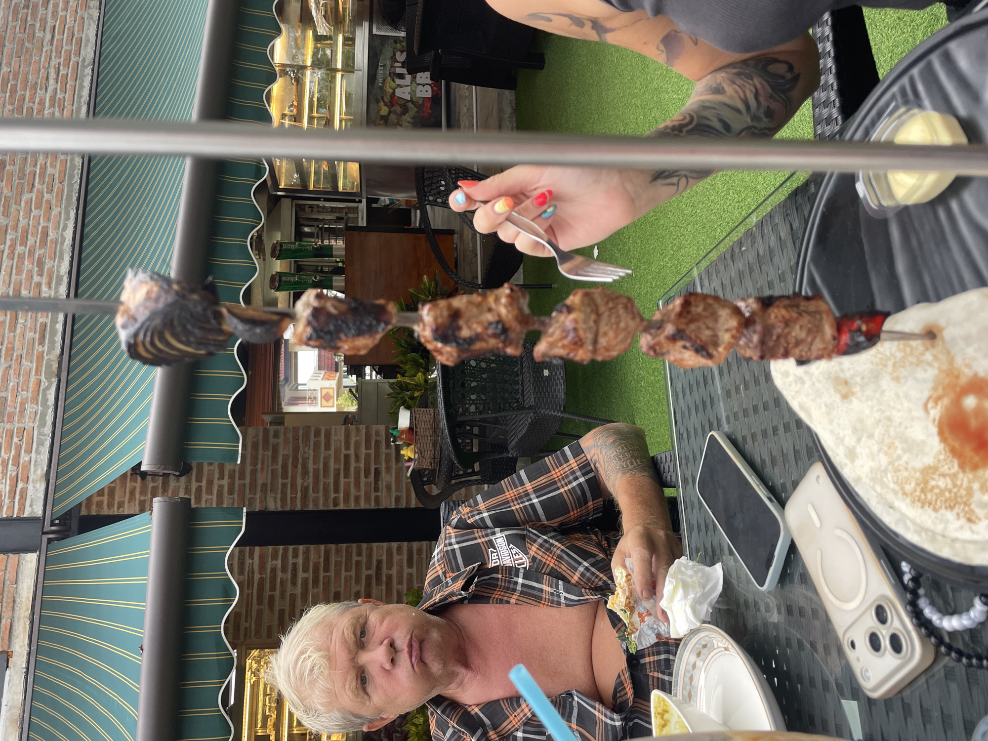 Captured outdoors in Choeng Thale, Thailand, the image presents an individual clad in an unbuttoned Harley-Davidson patterned shirt. The setting is a restaurant or eatery, implied by the presence of a table laden with food and dining implements. A metallic skewer, piercing through pieces of grilled meat, lies prominently across the scene. A female hand with multi-colored nail polish is seen using a fork to extract the meat from the skewer. A variety of items populate the tabletop: a partially eaten wrap, a small container of sauce, a plate, cutlery, two smartphones, and a drinking straw. Awnings displaying a stripe pattern offer shade, and a brick building is visible in the background, adding to the urban ambiance. The scene suggests a casual dining experience.