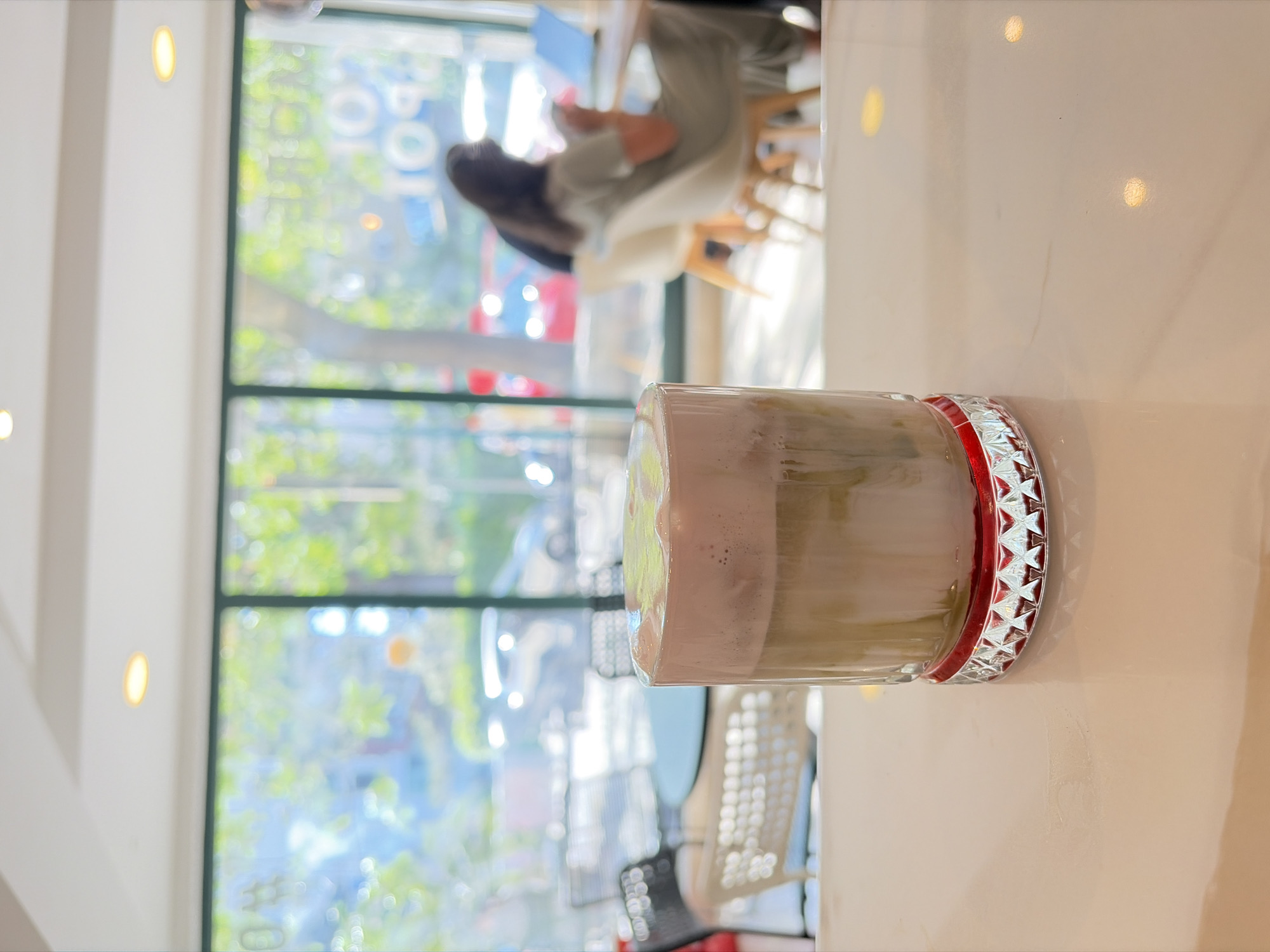 The image shows a close-up of a layered beverage in a clear glass with a decorative red and silver rim. The drink appears to have a creamy white layer on top, possibly foam, over a lighter brown or beige liquid. The glass is placed on a white, reflective surface, likely a table in a cafe or restaurant.
In the background, through a large window, a blurred scene of an outdoor area is visible. Trees with green foliage and hints of blue sky suggest it is daytime and likely pleasant weather. The interior of the establishment is also partially visible, with the suggestion of seating, including chairs and tables. A person, also blurred, is seen sitting at a table in the background, facing away from the camera and towards the window, possibly reading or working on a laptop, which is also faintly visible. The overall atmosphere seems relaxed and casual.
The image is taken from a slightly elevated angle, looking down at the drink. The lighting appears to be bright, with some light sources visible as orbs within the frame, hinting at indoor lighting. The scene is captured in Budva, Montenegro, though no specific landmarks are clearly identifiable. There is no discernible text visible in the image.