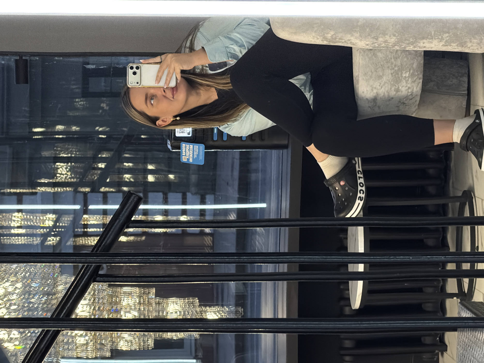 A woman is taking a picture of herself in a mirror in Moscow, Russia. She is wearing black leggings, a light blue shirt, white socks, and black Crocs. She is holding a white smartphone. In the background is a staircase with black rails and a chandelier reflected in the mirror. There is also what appears to be a sign with Russian text on it.