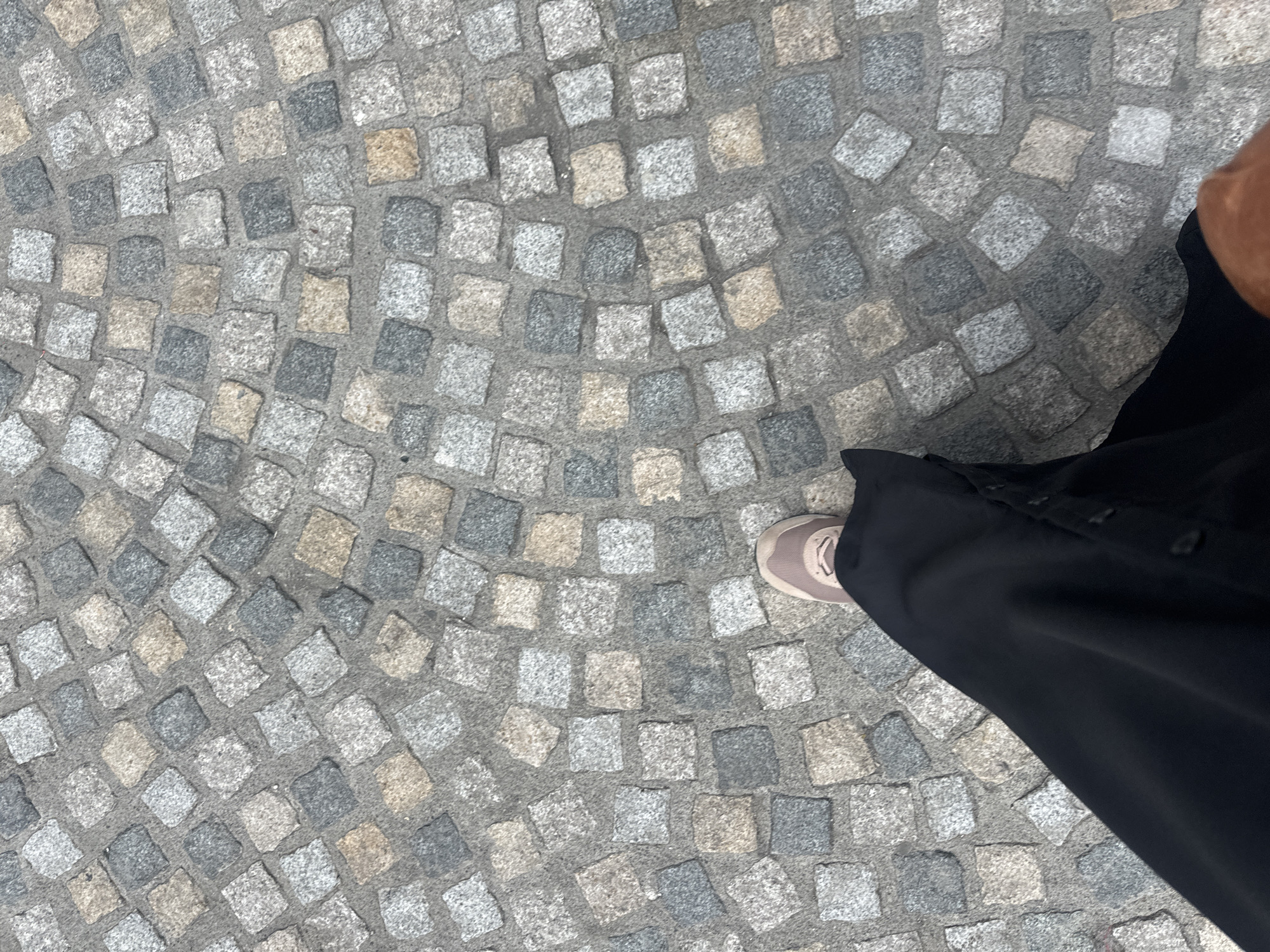A person is walking on a cobblestone street in Kilkenny, Ireland. The cobblestones are grey, tan, and dark grey squares laid in a fan pattern radiating outwards from the lower right of the frame. The person is wearing a black garment, possibly a long skirt or dress, and a light tan sneaker. The perspective is from above, looking down at the person's feet and the street.