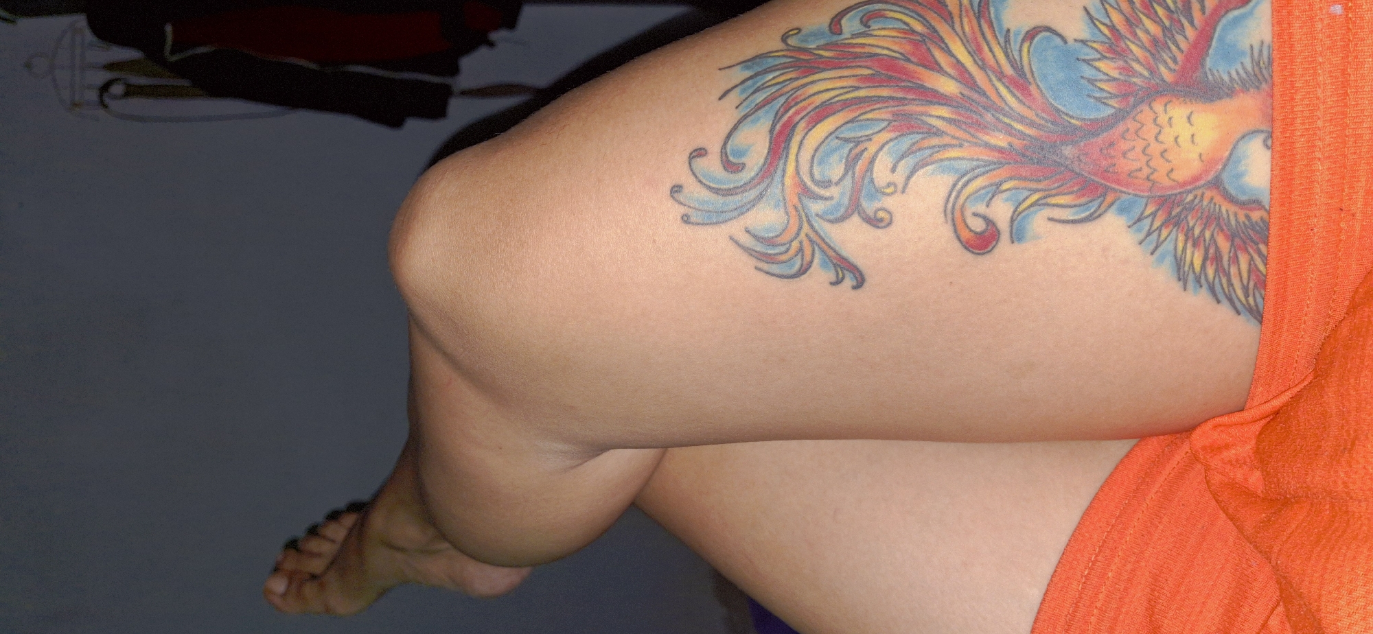 The image shows a close-up of a person's leg with a phoenix tattoo. The person is wearing an orange garment. The background is a dark blue wall with some clothing hanging on the right side. The image is taken in the city of Vallecito, Venezuela.