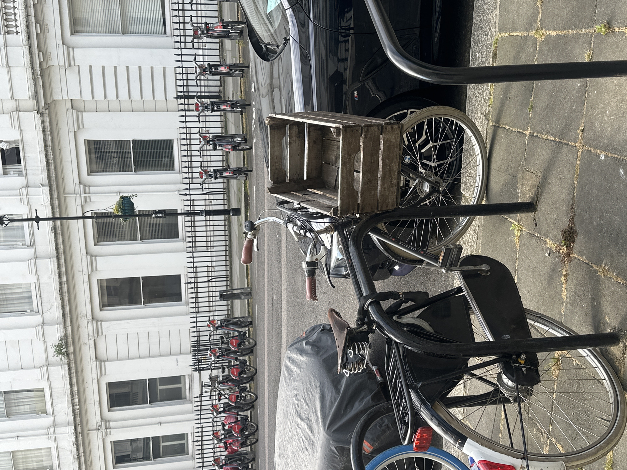 The image shows a street in London, United Kingdom. A black bicycle with a wooden crate attached to the front is parked on a paved sidewalk. The bicycle is leaning against a metal post, likely a bike rack. Behind the bicycle, there's a black car parked on the street. A black metal fence separates the sidewalk from a row of parked bicycles, all red and black. Behind the fence, there is a white building with multiple windows and architectural details. A lamppost with a hanging flower basket is visible. The overall scene suggests an urban environment, with a mix of transportation modes and architectural styles. The lighting indicates it's daytime.