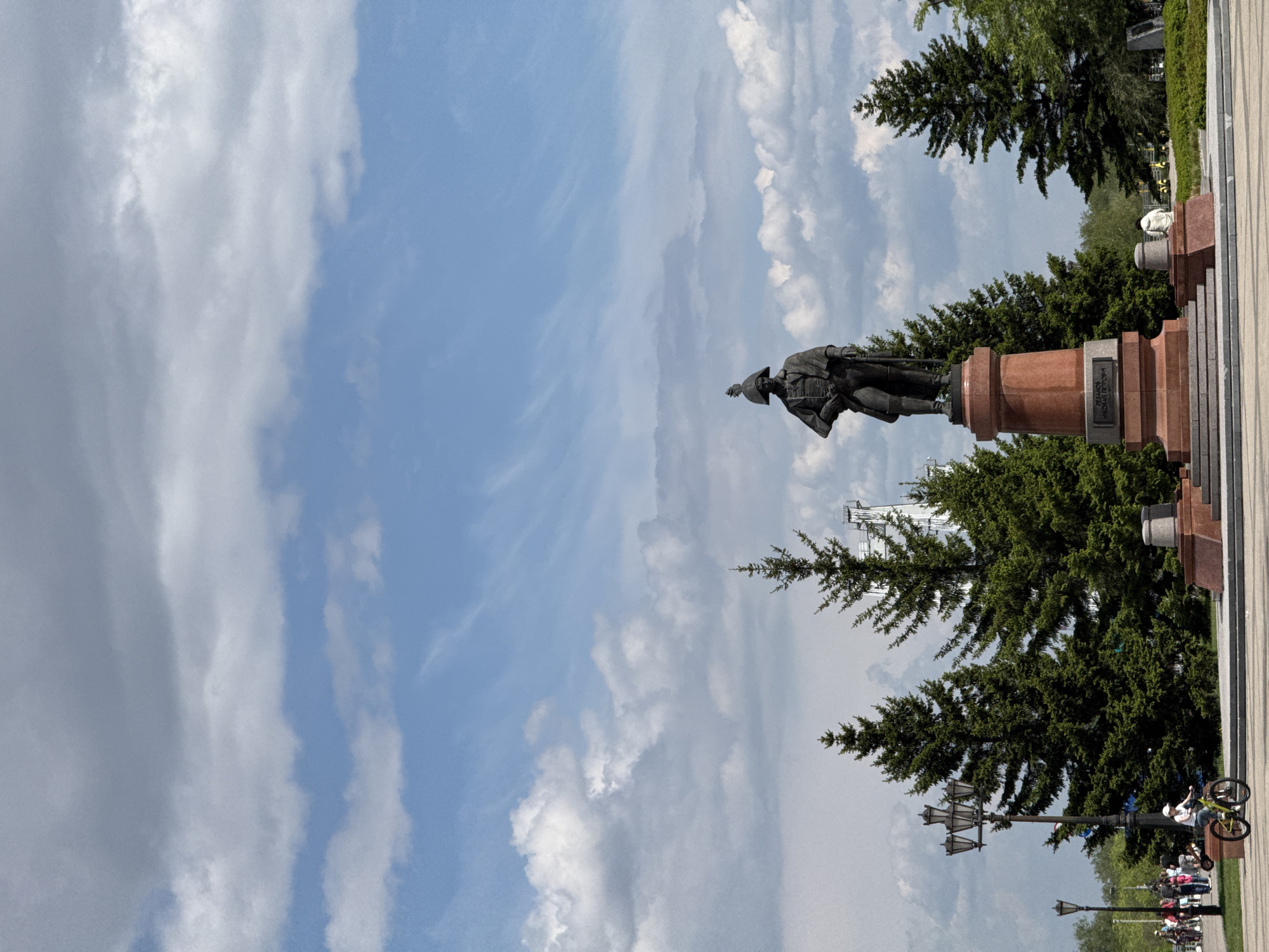 The image shows a statue of Andrey Dubensky, founder of Krasnoyarsk, Russia. The statue is positioned on a cylindrical pedestal atop a multi-tiered stone base. Behind the statue are trees and part of a white building. In the lower right, a sidewalk extends with lampposts and people. One individual is on a bicycle. The sky overhead is filled with clouds.