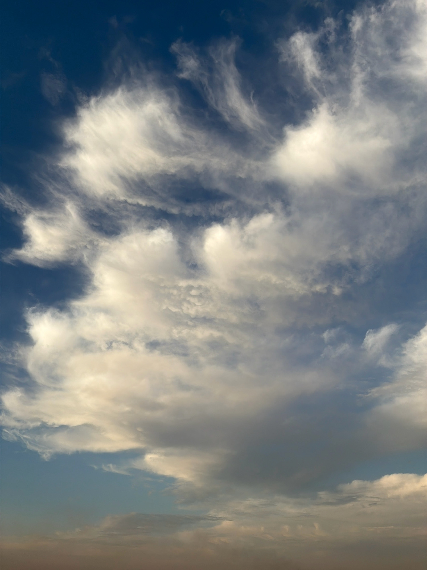 The image depicts a vast expanse of the sky above Astana, Kazakhstan, filled with a dynamic display of clouds. The deep blue of the upper atmosphere gradually transitions to lighter shades of blue and hints of warm hues closer to the horizon, suggesting either early morning or late afternoon.
The clouds are the primary subject, exhibiting a variety of forms. Wispy, feathery cirrus clouds streak across the upper portions of the image, their delicate tendrils catching the light. Below these, larger cumulus clouds are present, some appearing dense and voluminous with shaded undersides, while others are more scattered and translucent. There are also areas of darker, more uniform cloud cover, indicating potential changes in weather. The interplay of light and shadow across the cloud formations creates a sense of depth and movement within the scene.
There are no people, animals, buildings, or any terrestrial objects visible in this image, focusing solely on the atmospheric conditions above Astana. The environment is entirely the sky.