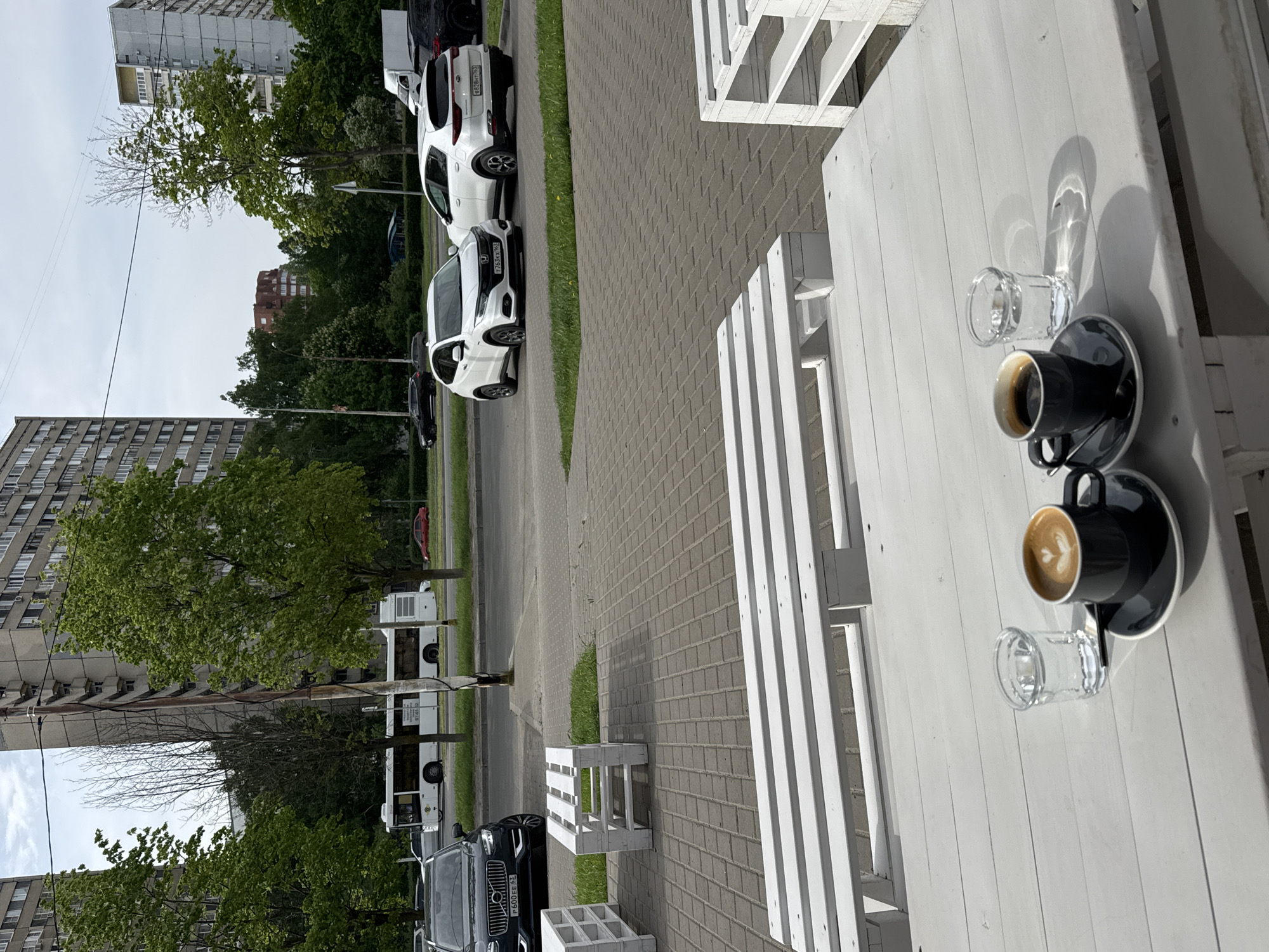 On a sunny day in an urban environment, two cups of coffee and two glasses of water are placed on a white wooden table, likely at an outdoor cafe. Cars are parked along the adjacent street. Buildings and trees make up the background.