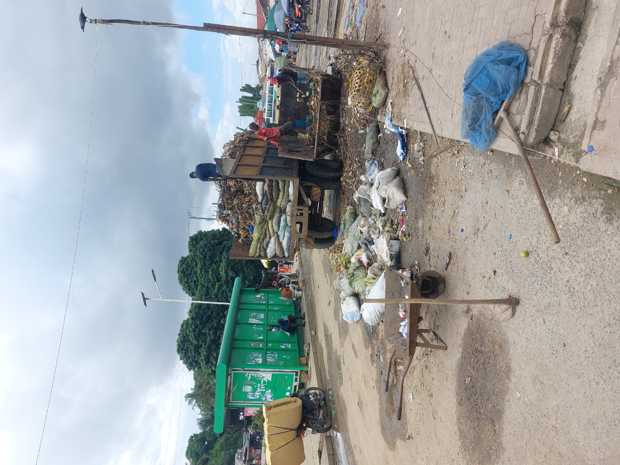 The image shows a scene in Dar es-Salaam, Tanzania, likely a public area with waste management challenges. A truck heavily loaded with various forms of refuse is present. People are working on the truck, seemingly sorting or loading waste. Beside the truck is a large green dumpster. Litter is strewn across the ground around the dumpster and the truck. A loaded motorcycle, possibly for transporting goods, is near the green dumpster. A wheelbarrow and various tools such as rakes are also visible amidst the trash. Power lines are above, and a bird is perched atop a light pole. The sky is overcast. The area appears to be an informal market or commercial area, with people in the background and structures nearby. The general impression is one of poor sanitation and waste management in a busy urban environment.