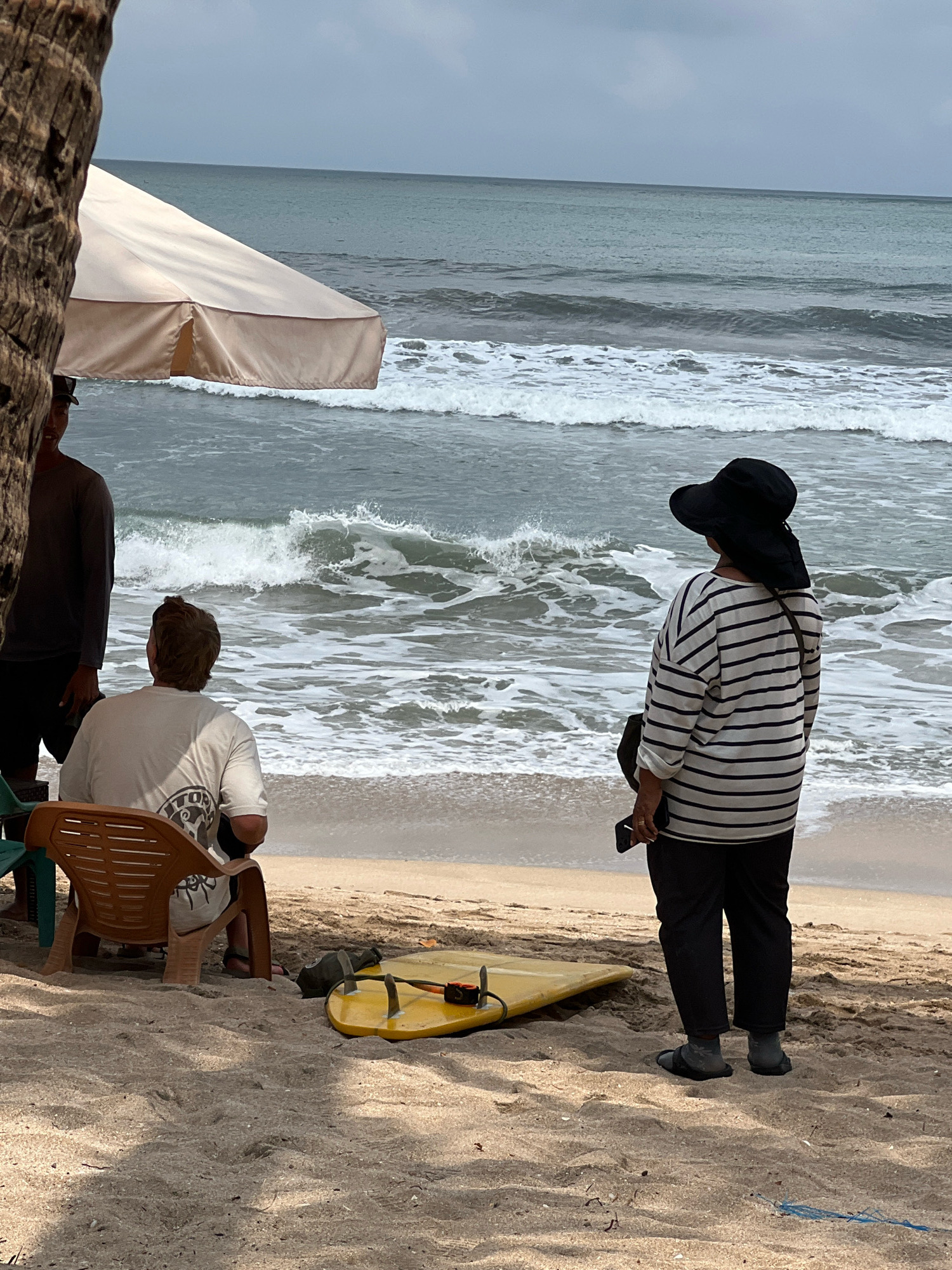 An overcast day at Kuta Beach in Kuta, Indonesia, shows three individuals on the sandy shore near the ocean. On the left, partially obscured by a palm tree and a light-colored beach umbrella, a person wearing a gray long-sleeved shirt and dark shorts stands. In the center, a person with short brown hair sits in a brown plastic chair, facing away from the viewer and towards the ocean, wearing a light-colored t-shirt with a graphic on the back. To the right, a person wearing a wide-brimmed black hat, a horizontally striped long-sleeved shirt, and dark pants stands facing the ocean, holding a black object, possibly a phone, in their right hand. A yellow surfboard lies on the sand in front of the person seated in the chair. The ocean exhibits moderate waves with white foam breaking near the shore. The sand is visible in the foreground, with some shadows cast from unseen objects. The sky is uniformly gray.