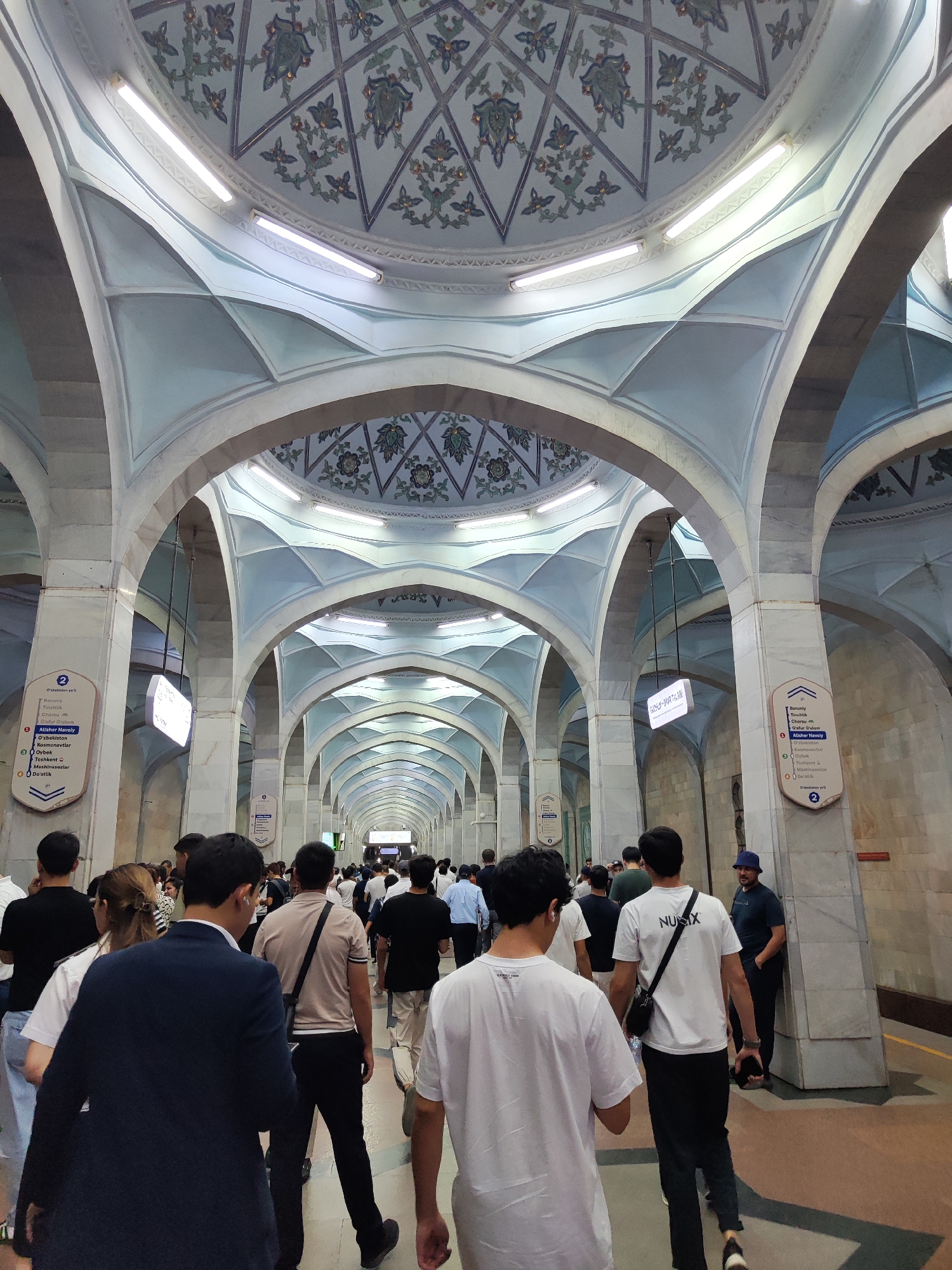 The image depicts the interior of a subway station in Tashkent, Uzbekistan, characterized by its ornate architecture. The station features a high, vaulted ceiling adorned with intricate floral patterns in shades of blue and green, set against a white background. Arched passageways with marble-clad columns line the corridor, creating a sense of depth and grandeur. Fluorescent lighting fixtures are embedded in the ceiling.
A crowd of people, predominantly adults dressed in casual clothing, are seen walking through the station. Many are facing away from the camera, indicating movement in a particular direction. The floor is a light-colored tile with yellow lines marking walkways. Signs with Cyrillic and Latin script are mounted on the pillars, providing station information and directional cues. One sign, featuring a blue shield with the number '2', lists various station names, including "Alisher Navoi". The overall atmosphere suggests a busy urban transit environment.